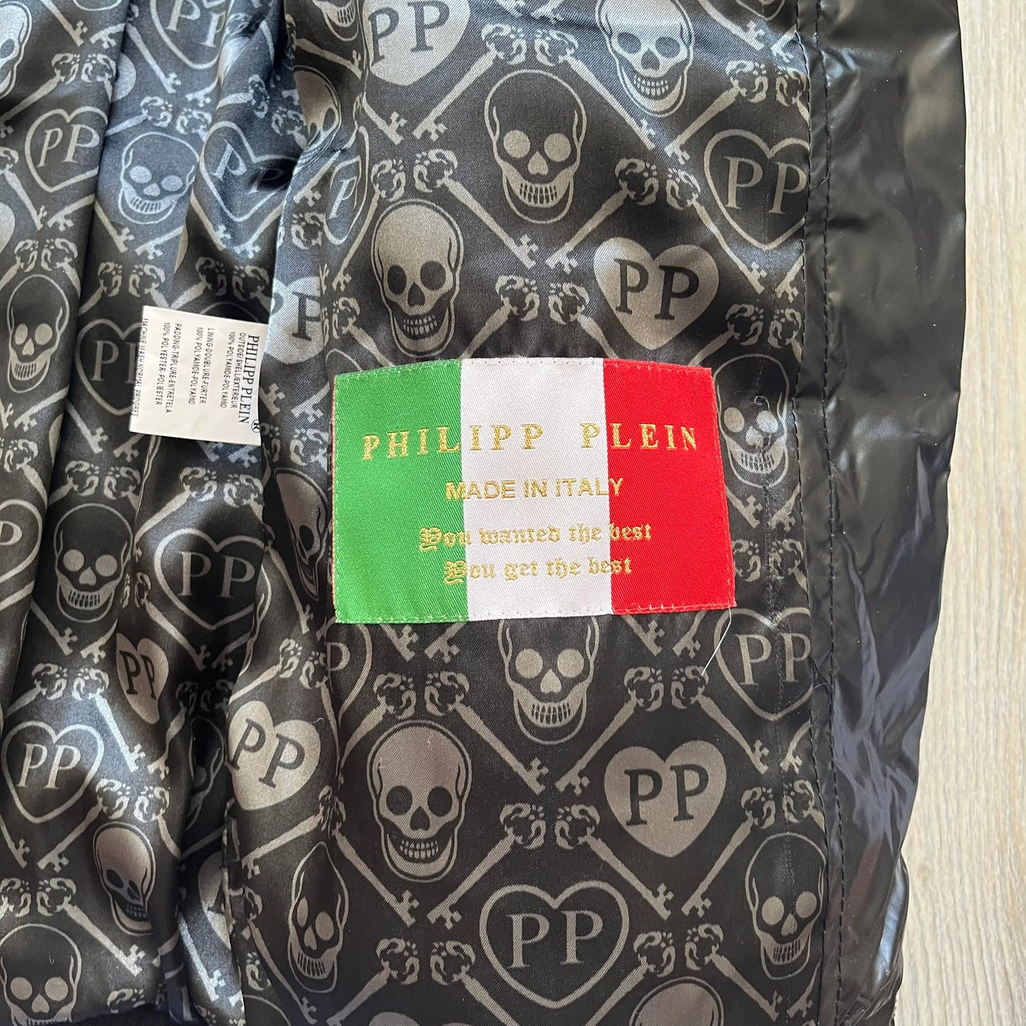 Philipp Plein 2010's Black Waxed Puffer Jacket is a black puffer jacket with a removable fur-trimmed hood attached by zipper. The jacket features heavy silver-tone hardware throughout, including large zipper pulls shaped as crosses with a skull in the center. The right chest has a crystal “P” patch set, while the left chest features two zip pockets. The hood drawstrings are finished with silver skull tips marked with a “P.”

One shoulder is finished with metal Philipp Plein logo plaque. The interior lining 