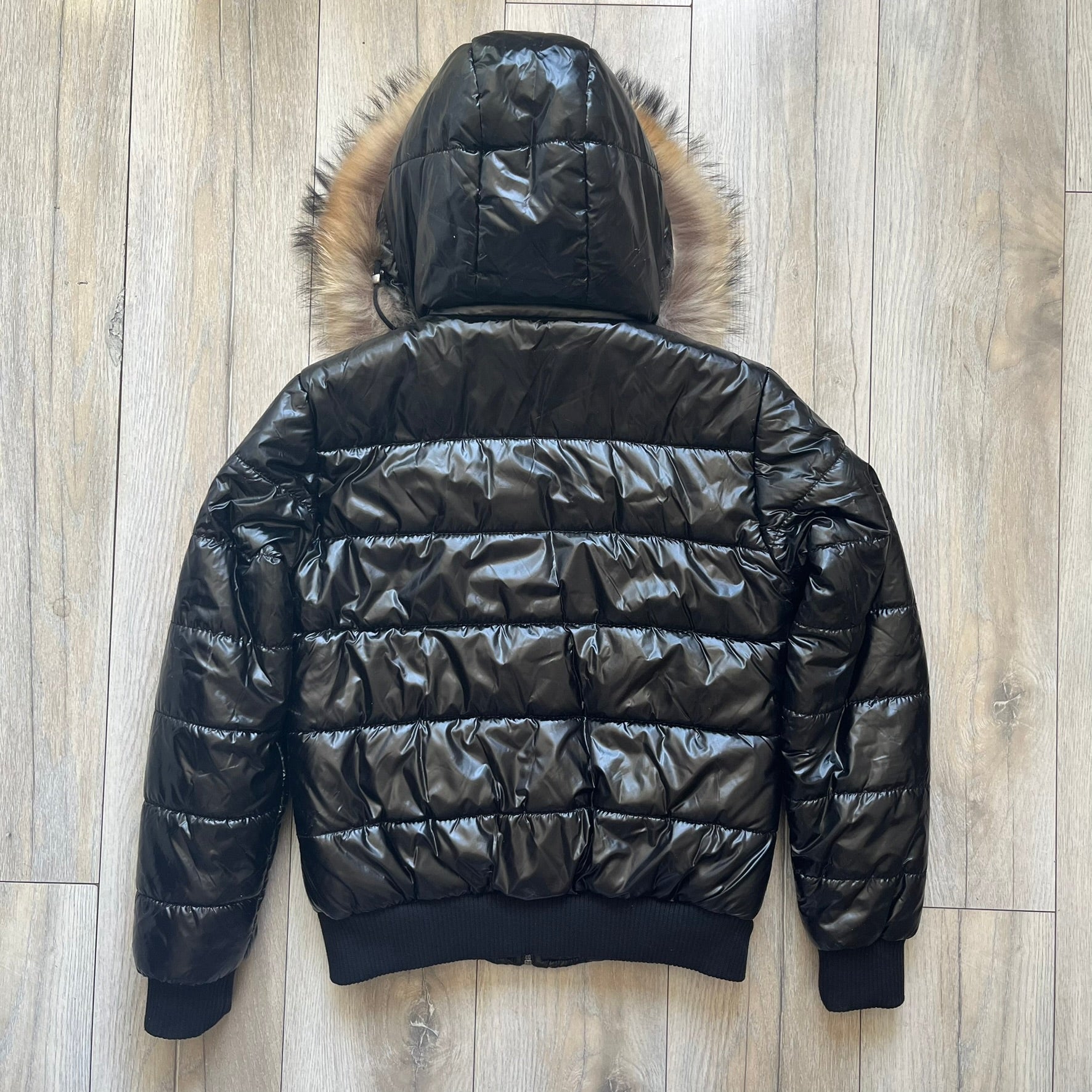 Philipp Plein 2010's Black Waxed Puffer Jacket is a black puffer jacket with a removable fur-trimmed hood attached by zipper. The jacket features heavy silver-tone hardware throughout, including large zipper pulls shaped as crosses with a skull in the center. The right chest has a crystal “P” patch set, while the left chest features two zip pockets. The hood drawstrings are finished with silver skull tips marked with a “P.”

One shoulder is finished with metal Philipp Plein logo plaque. The interior lining 