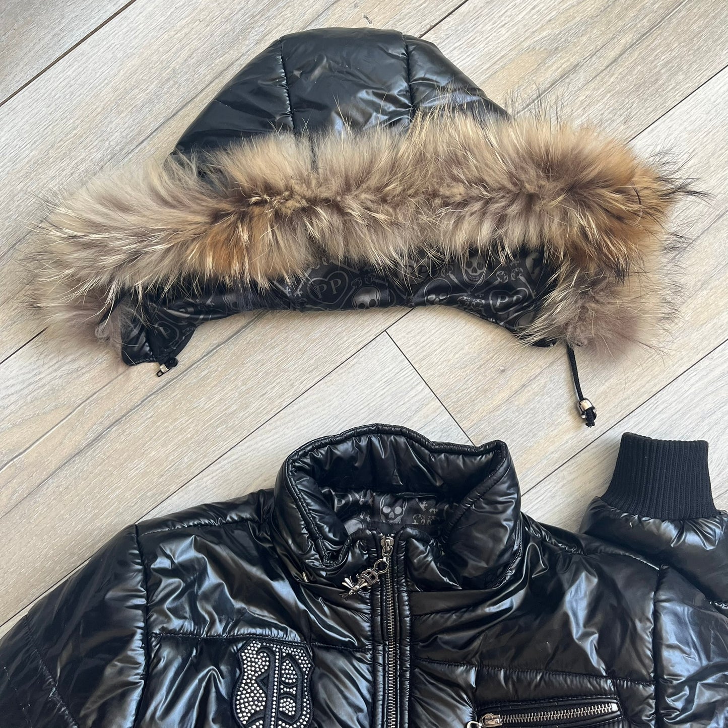 Philipp Plein 2010's Black Waxed Puffer Jacket is a black puffer jacket with a removable fur-trimmed hood attached by zipper. The jacket features heavy silver-tone hardware throughout, including large zipper pulls shaped as crosses with a skull in the center. The right chest has a crystal “P” patch set, while the left chest features two zip pockets. The hood drawstrings are finished with silver skull tips marked with a “P.”

One shoulder is finished with metal Philipp Plein logo plaque. The interior lining 