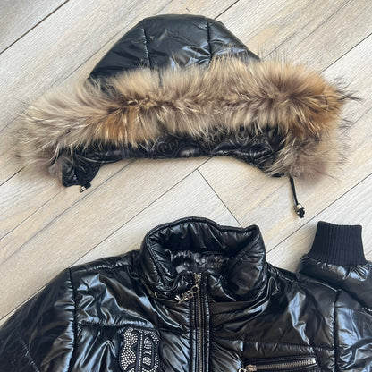 Philipp Plein 2010's Black Waxed Puffer Jacket is a black puffer jacket with a removable fur-trimmed hood attached by zipper. The jacket features heavy silver-tone hardware throughout, including large zipper pulls shaped as crosses with a skull in the center. The right chest has a crystal “P” patch set, while the left chest features two zip pockets. The hood drawstrings are finished with silver skull tips marked with a “P.”

One shoulder is finished with metal Philipp Plein logo plaque. The interior lining 