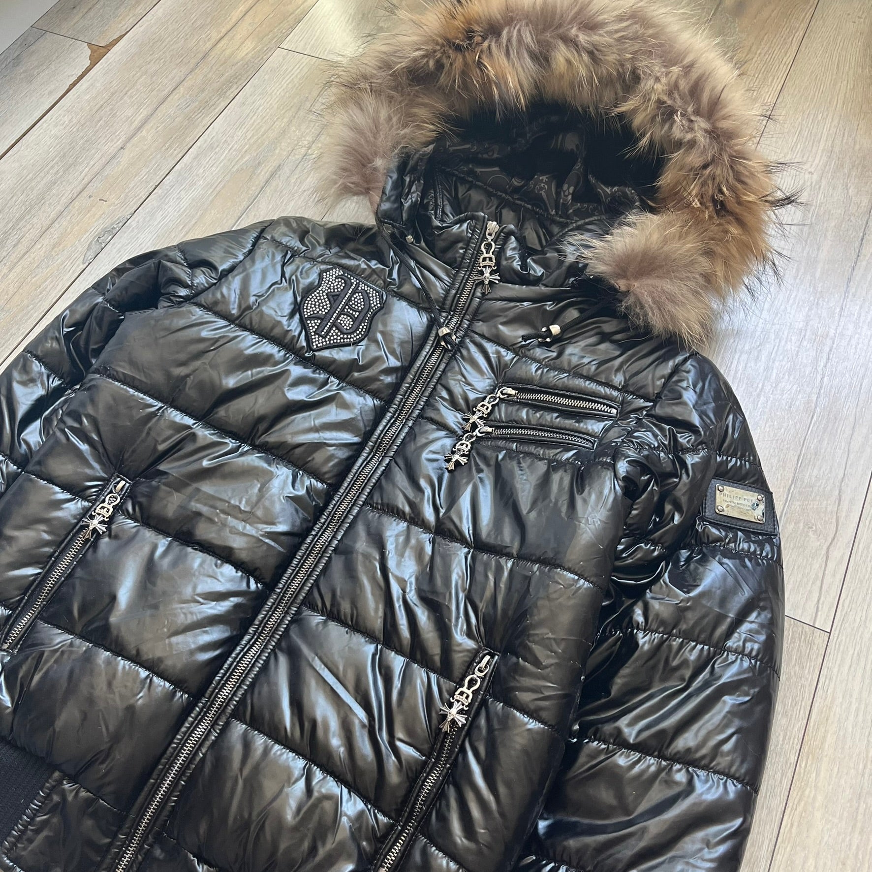 Philipp Plein 2010's Black Waxed Puffer Jacket is a black puffer jacket with a removable fur-trimmed hood attached by zipper. The jacket features heavy silver-tone hardware throughout, including large zipper pulls shaped as crosses with a skull in the center. The right chest has a crystal “P” patch set, while the left chest features two zip pockets. The hood drawstrings are finished with silver skull tips marked with a “P.”

One shoulder is finished with metal Philipp Plein logo plaque. The interior lining 