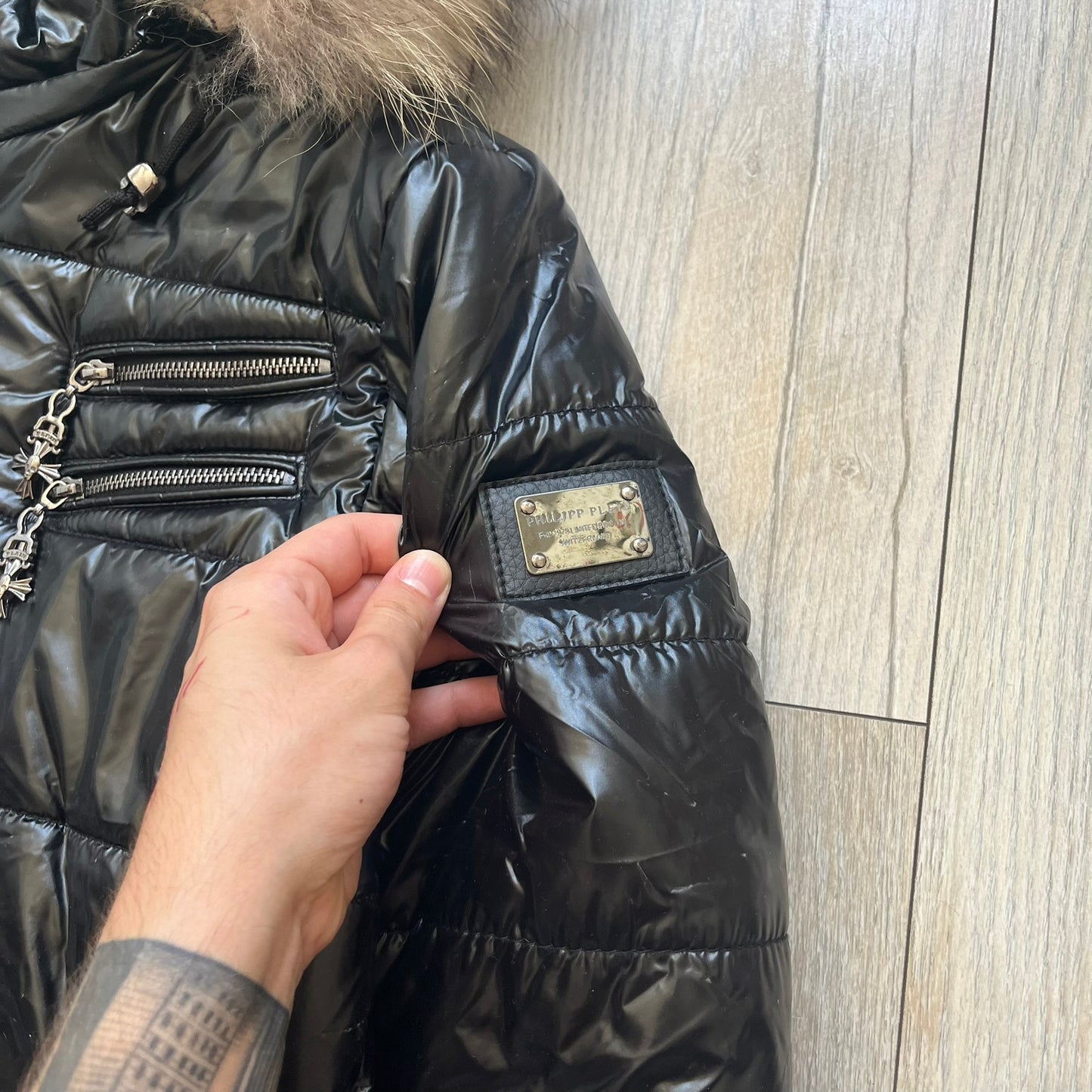 Philipp Plein 2010's Black Waxed Puffer Jacket is a black puffer jacket with a removable fur-trimmed hood attached by zipper. The jacket features heavy silver-tone hardware throughout, including large zipper pulls shaped as crosses with a skull in the center. The right chest has a crystal “P” patch set, while the left chest features two zip pockets. The hood drawstrings are finished with silver skull tips marked with a “P.”

One shoulder is finished with metal Philipp Plein logo plaque. The interior lining 