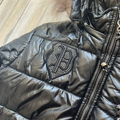 Philipp Plein 2010's Black Waxed Puffer Jacket is a black puffer jacket with a removable fur-trimmed hood attached by zipper. The jacket features heavy silver-tone hardware throughout, including large zipper pulls shaped as crosses with a skull in the center. The right chest has a crystal “P” patch set, while the left chest features two zip pockets. The hood drawstrings are finished with silver skull tips marked with a “P.”

One shoulder is finished with metal Philipp Plein logo plaque. The interior lining 