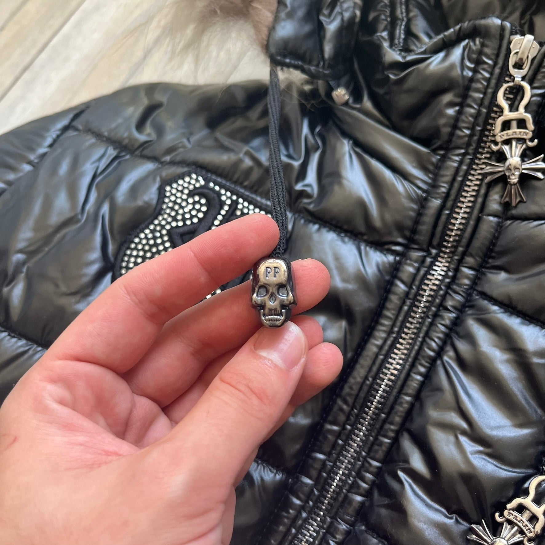 Philipp Plein 2010's Black Waxed Puffer Jacket is a black puffer jacket with a removable fur-trimmed hood attached by zipper. The jacket features heavy silver-tone hardware throughout, including large zipper pulls shaped as crosses with a skull in the center. The right chest has a crystal “P” patch set, while the left chest features two zip pockets. The hood drawstrings are finished with silver skull tips marked with a “P.”

One shoulder is finished with metal Philipp Plein logo plaque. The interior lining 