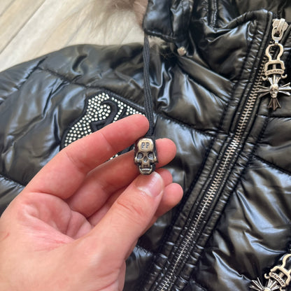 Philipp Plein 2010's Black Waxed Puffer Jacket is a black puffer jacket with a removable fur-trimmed hood attached by zipper. The jacket features heavy silver-tone hardware throughout, including large zipper pulls shaped as crosses with a skull in the center. The right chest has a crystal “P” patch set, while the left chest features two zip pockets. The hood drawstrings are finished with silver skull tips marked with a “P.”

One shoulder is finished with metal Philipp Plein logo plaque. The interior lining 