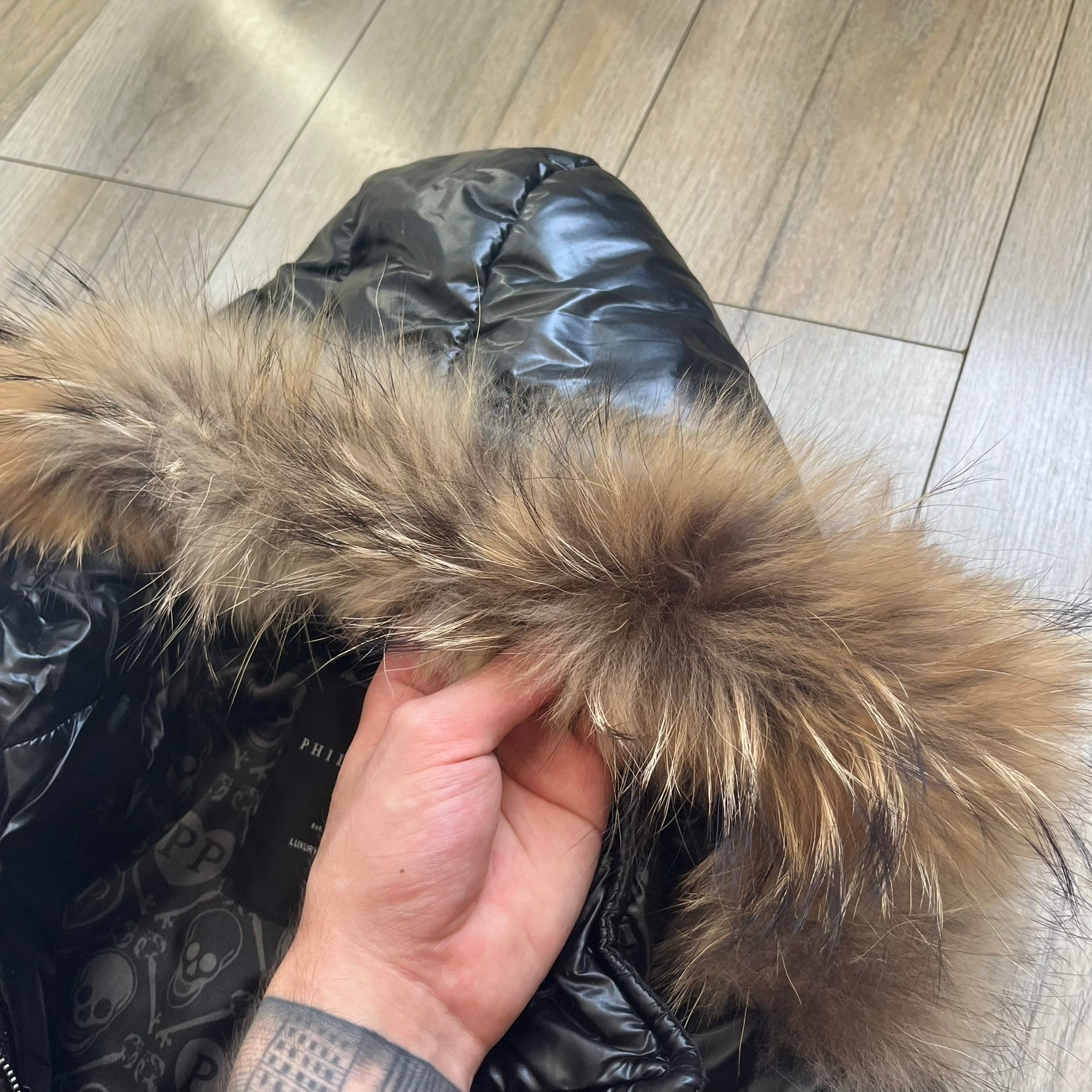 Philipp Plein 2010's Black Waxed Puffer Jacket is a black puffer jacket with a removable fur-trimmed hood attached by zipper. The jacket features heavy silver-tone hardware throughout, including large zipper pulls shaped as crosses with a skull in the center. The right chest has a crystal “P” patch set, while the left chest features two zip pockets. The hood drawstrings are finished with silver skull tips marked with a “P.”

One shoulder is finished with metal Philipp Plein logo plaque. The interior lining 