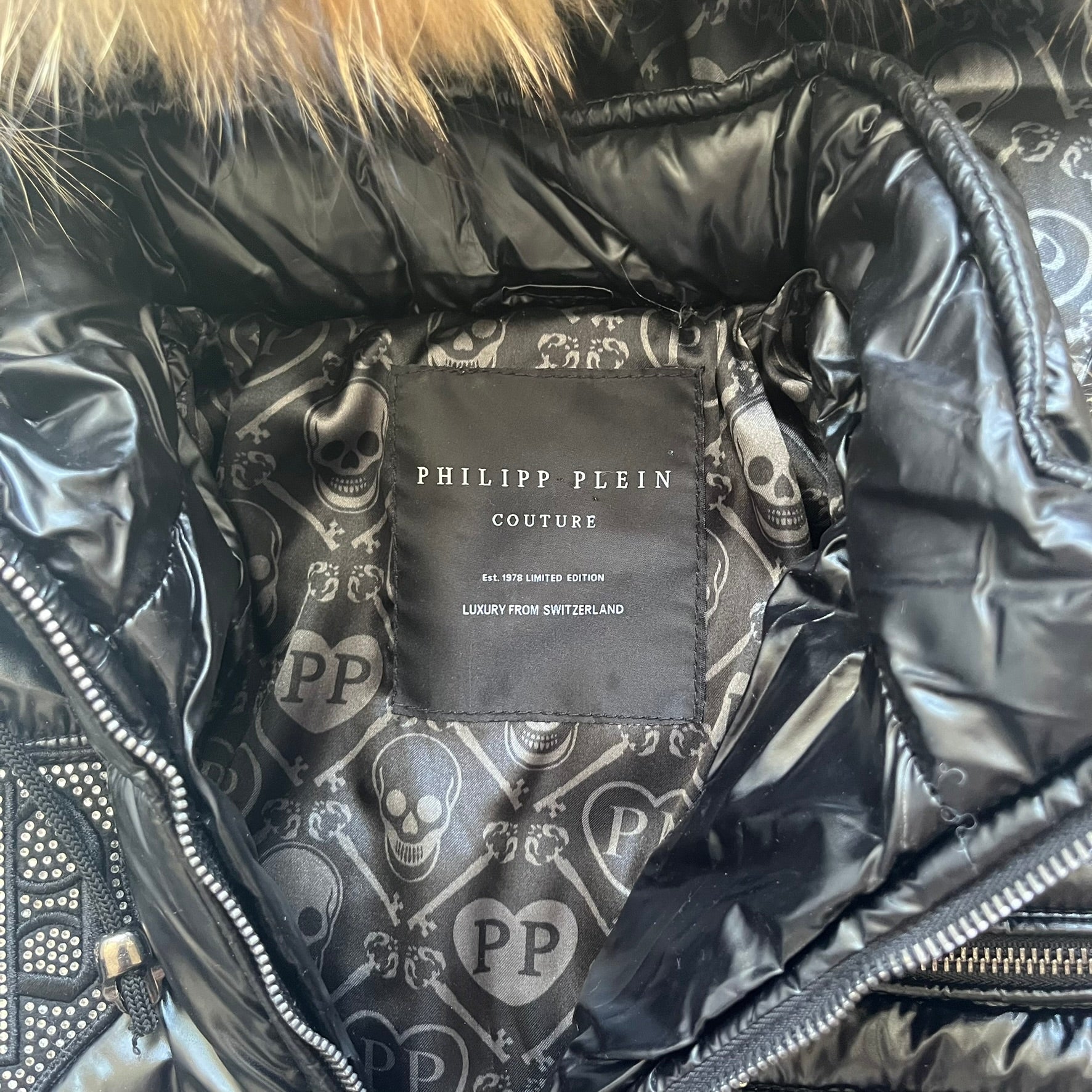 Philipp Plein 2010's Black Waxed Puffer Jacket is a black puffer jacket with a removable fur-trimmed hood attached by zipper. The jacket features heavy silver-tone hardware throughout, including large zipper pulls shaped as crosses with a skull in the center. The right chest has a crystal “P” patch set, while the left chest features two zip pockets. The hood drawstrings are finished with silver skull tips marked with a “P.”

One shoulder is finished with metal Philipp Plein logo plaque. The interior lining 