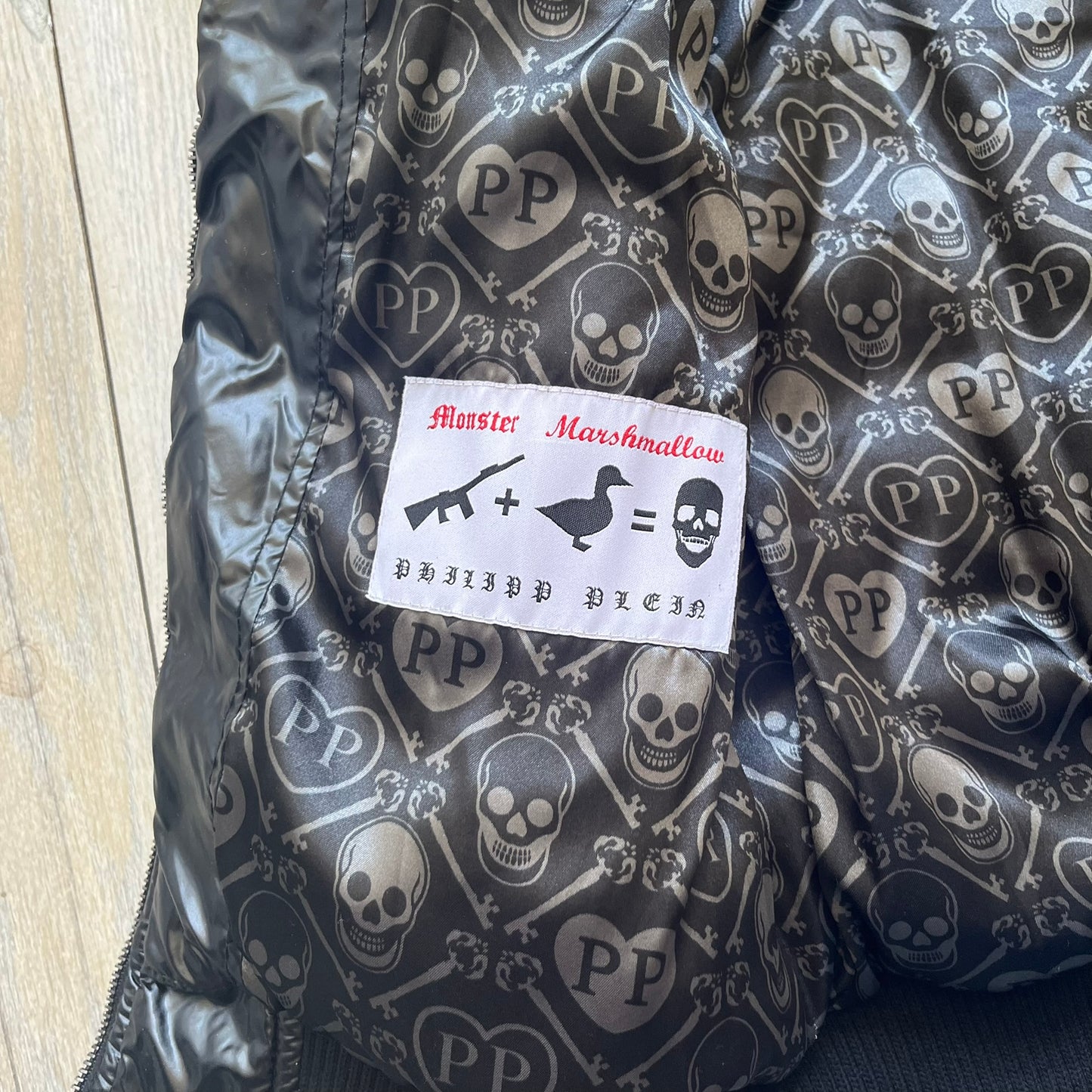 Philipp Plein 2010's Black Waxed Puffer Jacket is a black puffer jacket with a removable fur-trimmed hood attached by zipper. The jacket features heavy silver-tone hardware throughout, including large zipper pulls shaped as crosses with a skull in the center. The right chest has a crystal “P” patch set, while the left chest features two zip pockets. The hood drawstrings are finished with silver skull tips marked with a “P.”

One shoulder is finished with metal Philipp Plein logo plaque. The interior lining 