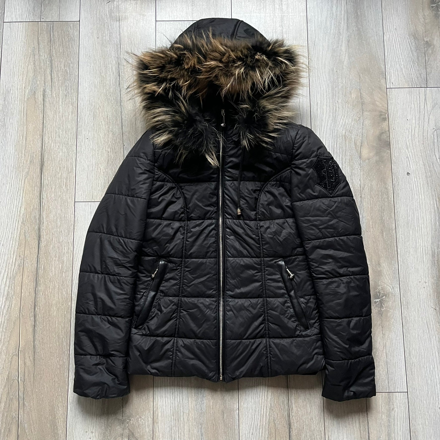 Philipp Plein 2010's Fur Hood Puffer Jacket is a black puffer jacket with a removable fur-trimmed hood attached by zipper. The jacket features heavy silver-tone hardware throughout, including 3 large zipper pulls used on the front pockets, main zipper and one drawstring ending. The right arm has a black “P” patch set with crystals.
