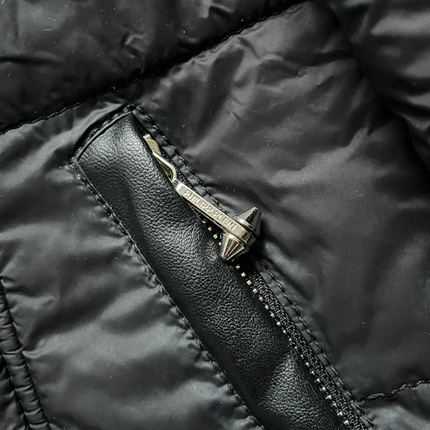Philipp Plein 2010's Fur Hood Puffer Jacket is a black puffer jacket with a removable fur-trimmed hood attached by zipper. The jacket features heavy silver-tone hardware throughout, including 3 large zipper pulls used on the front pockets, main zipper and one drawstring ending. The right arm has a black “P” patch set with crystals.
