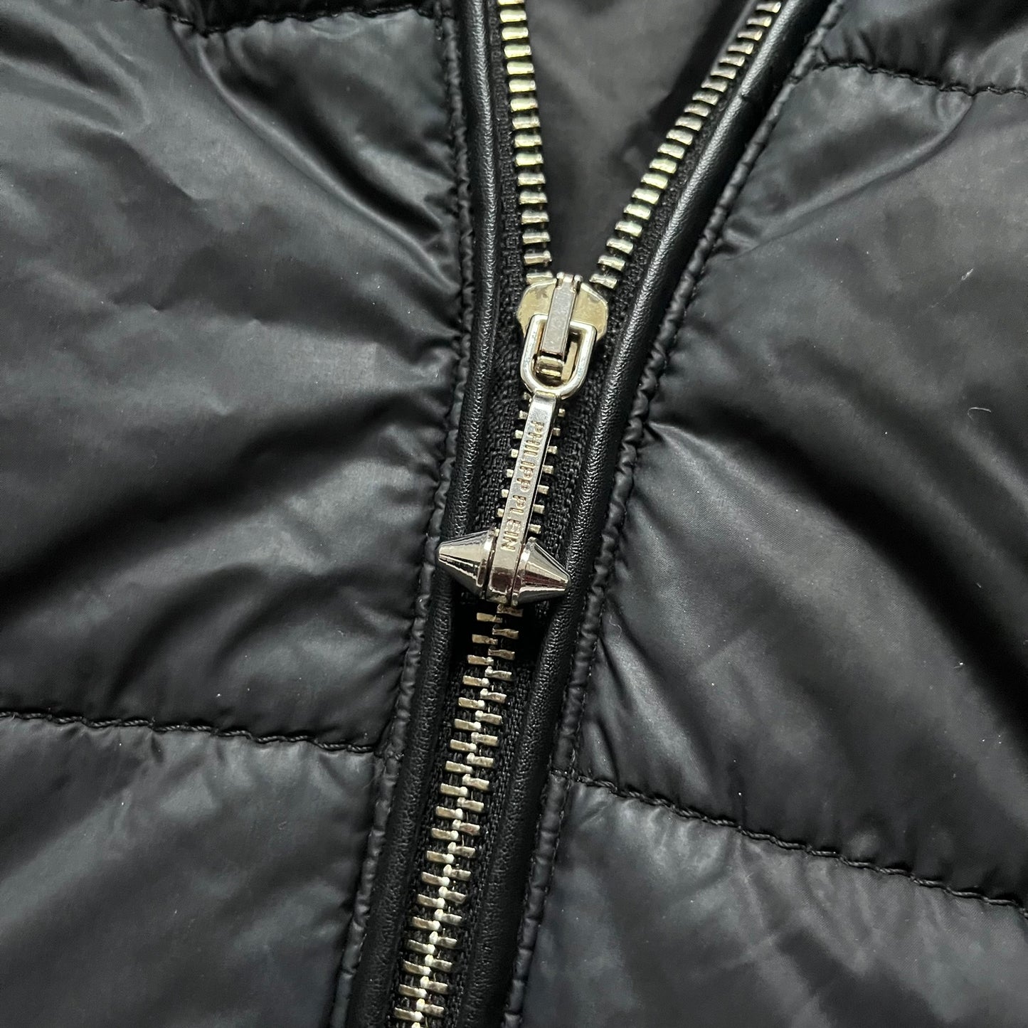 Philipp Plein 2010's Fur Hood Puffer Jacket is a black puffer jacket with a removable fur-trimmed hood attached by zipper. The jacket features heavy silver-tone hardware throughout, including 3 large zipper pulls used on the front pockets, main zipper and one drawstring ending. The right arm has a black “P” patch set with crystals.