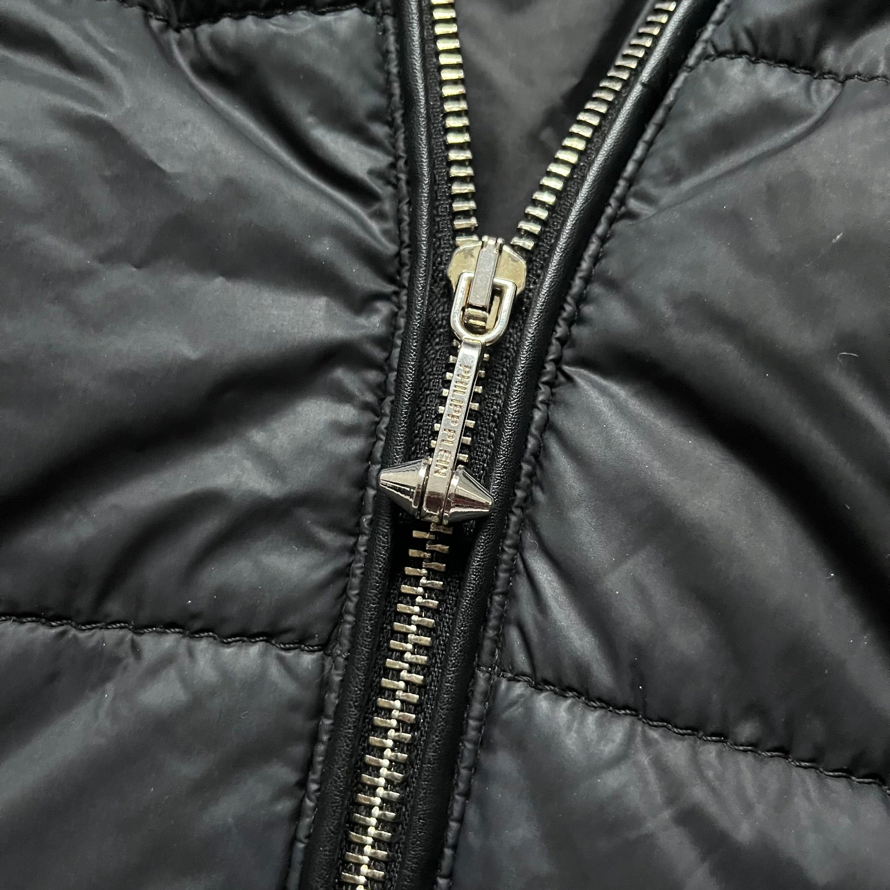 Philipp Plein 2010's Fur Hood Puffer Jacket is a black puffer jacket with a removable fur-trimmed hood attached by zipper. The jacket features heavy silver-tone hardware throughout, including 3 large zipper pulls used on the front pockets, main zipper and one drawstring ending. The right arm has a black “P” patch set with crystals.