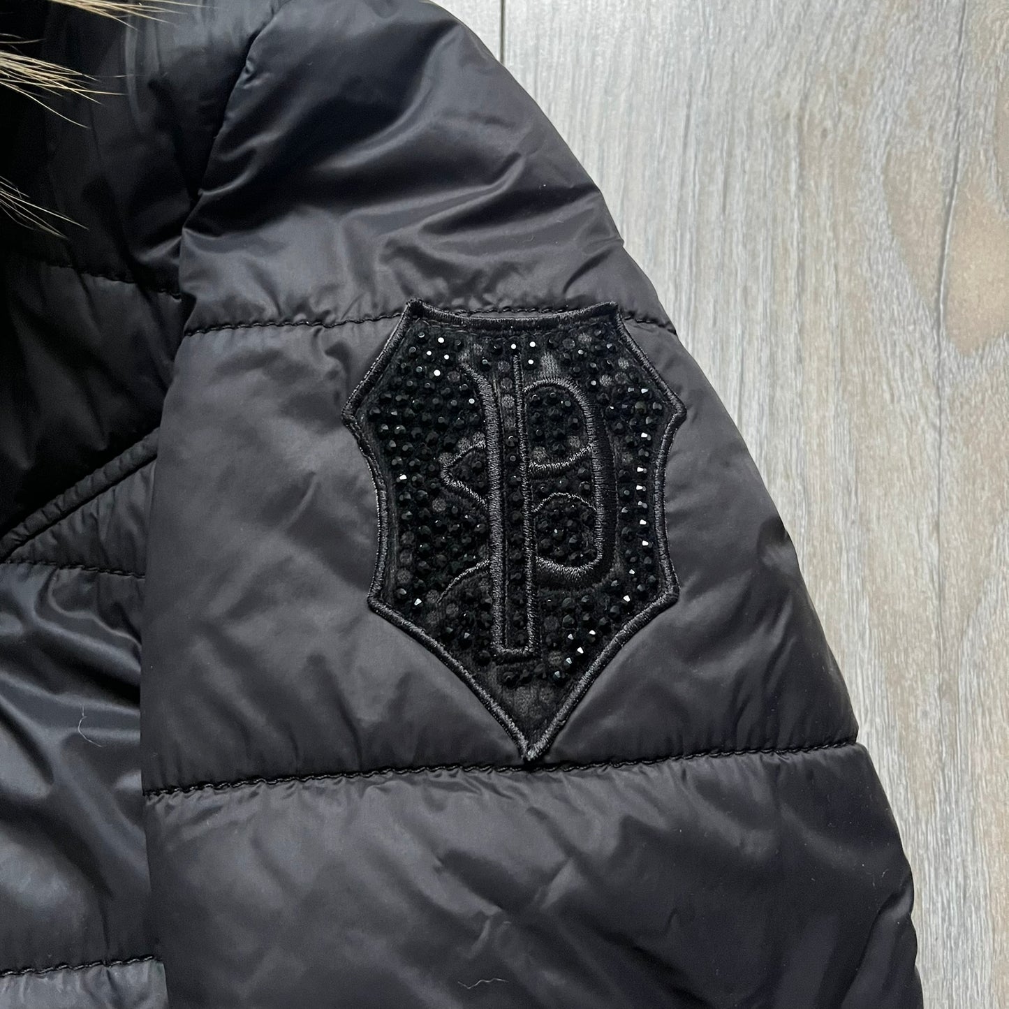Philipp Plein 2010's Fur Hood Puffer Jacket is a black puffer jacket with a removable fur-trimmed hood attached by zipper. The jacket features heavy silver-tone hardware throughout, including 3 large zipper pulls used on the front pockets, main zipper and one drawstring ending. The right arm has a black “P” patch set with crystals.