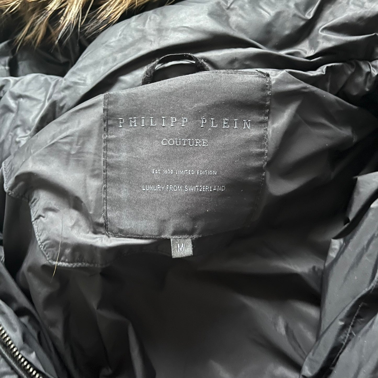 Philipp Plein 2010's Fur Hood Puffer Jacket is a black puffer jacket with a removable fur-trimmed hood attached by zipper. The jacket features heavy silver-tone hardware throughout, including 3 large zipper pulls used on the front pockets, main zipper and one drawstring ending. The right arm has a black “P” patch set with crystals.