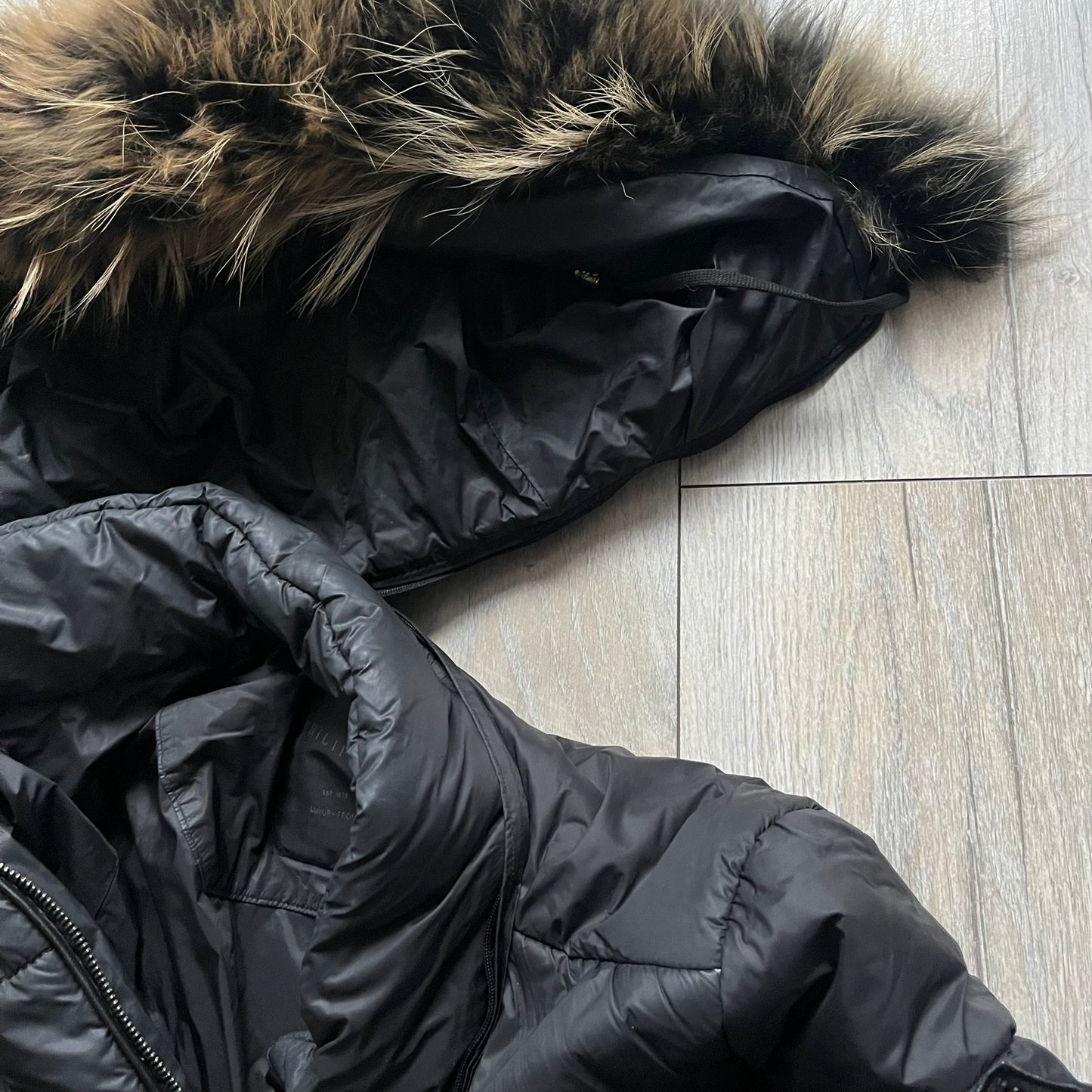 Philipp Plein 2010's Fur Hood Puffer Jacket is a black puffer jacket with a removable fur-trimmed hood attached by zipper. The jacket features heavy silver-tone hardware throughout, including 3 large zipper pulls used on the front pockets, main zipper and one drawstring ending. The right arm has a black “P” patch set with crystals.