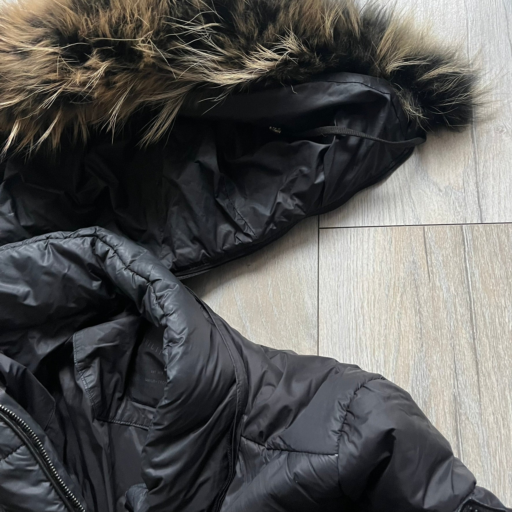 Philipp Plein 2010's Fur Hood Puffer Jacket is a black puffer jacket with a removable fur-trimmed hood attached by zipper. The jacket features heavy silver-tone hardware throughout, including 3 large zipper pulls used on the front pockets, main zipper and one drawstring ending. The right arm has a black “P” patch set with crystals.
