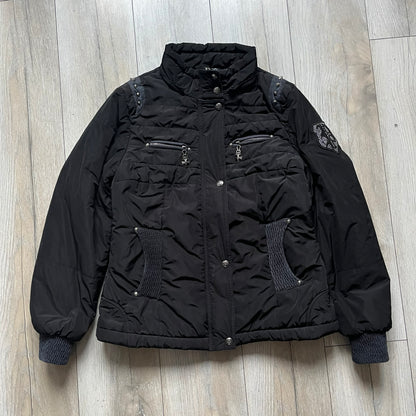 Philipp Plein 2010's No Hood Puffer Jacket is a black puffer jacket, with silver-tone hardware throughout including large zipper pulls shaped as crosses with a skull in the center used on the pockets, buttons with PP Skulls and metal "spikes" on the shoulders. The right arm has a white/clear “P” patch set with crystals. The interior lining has an all-over print featuring keys, skulls, and PP logos.