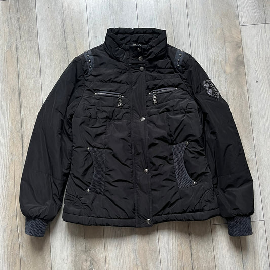 Philipp Plein 2010's No Hood Puffer Jacket is a black puffer jacket, with silver-tone hardware throughout including large zipper pulls shaped as crosses with a skull in the center used on the pockets, buttons with PP Skulls and metal "spikes" on the shoulders. The right arm has a white/clear “P” patch set with crystals. The interior lining has an all-over print featuring keys, skulls, and PP logos.