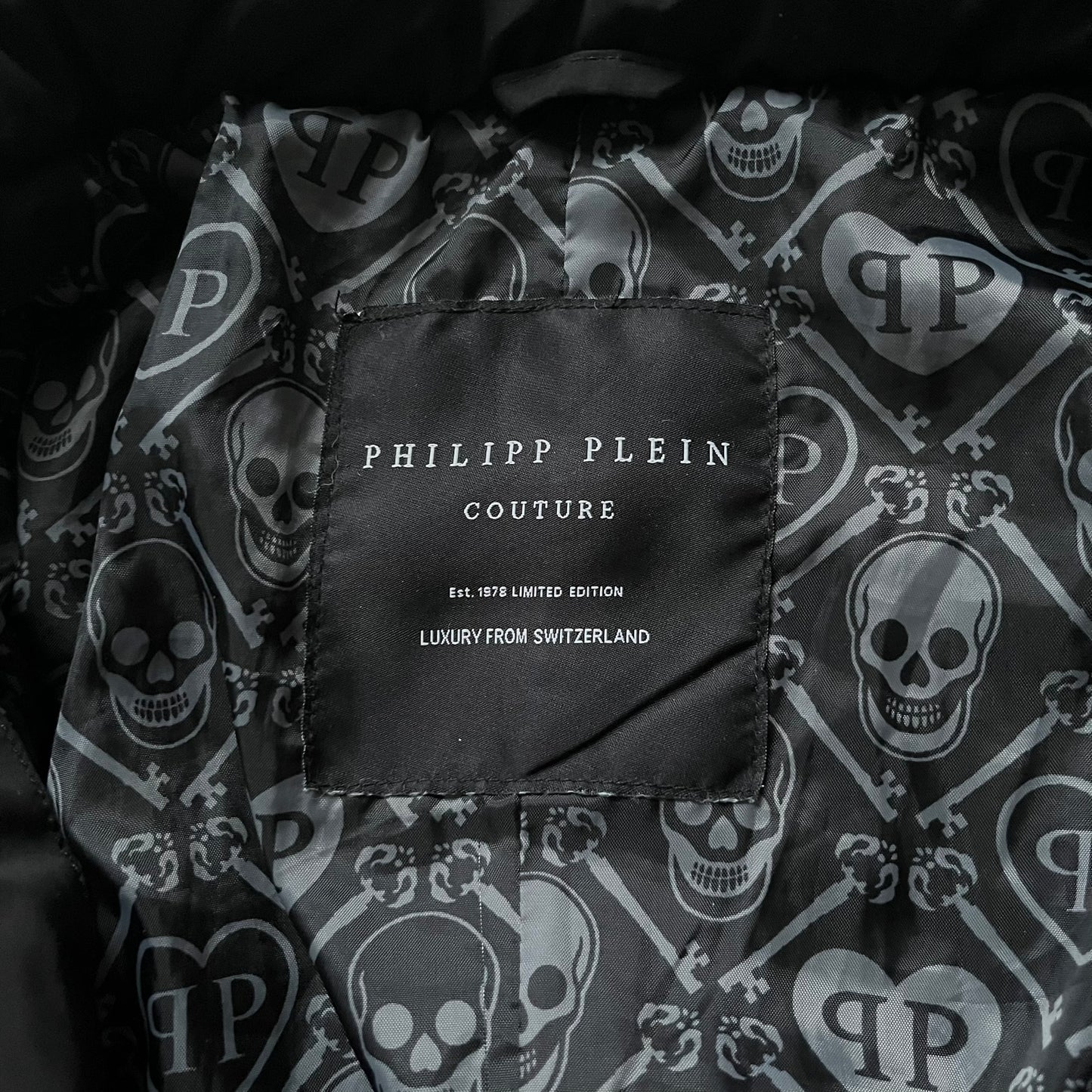 Philipp Plein 2010's No Hood Puffer Jacket is a black puffer jacket, with silver-tone hardware throughout including large zipper pulls shaped as crosses with a skull in the center used on the pockets, buttons with PP Skulls and metal "spikes" on the shoulders. The right arm has a white/clear “P” patch set with crystals. The interior lining has an all-over print featuring keys, skulls, and PP logos.