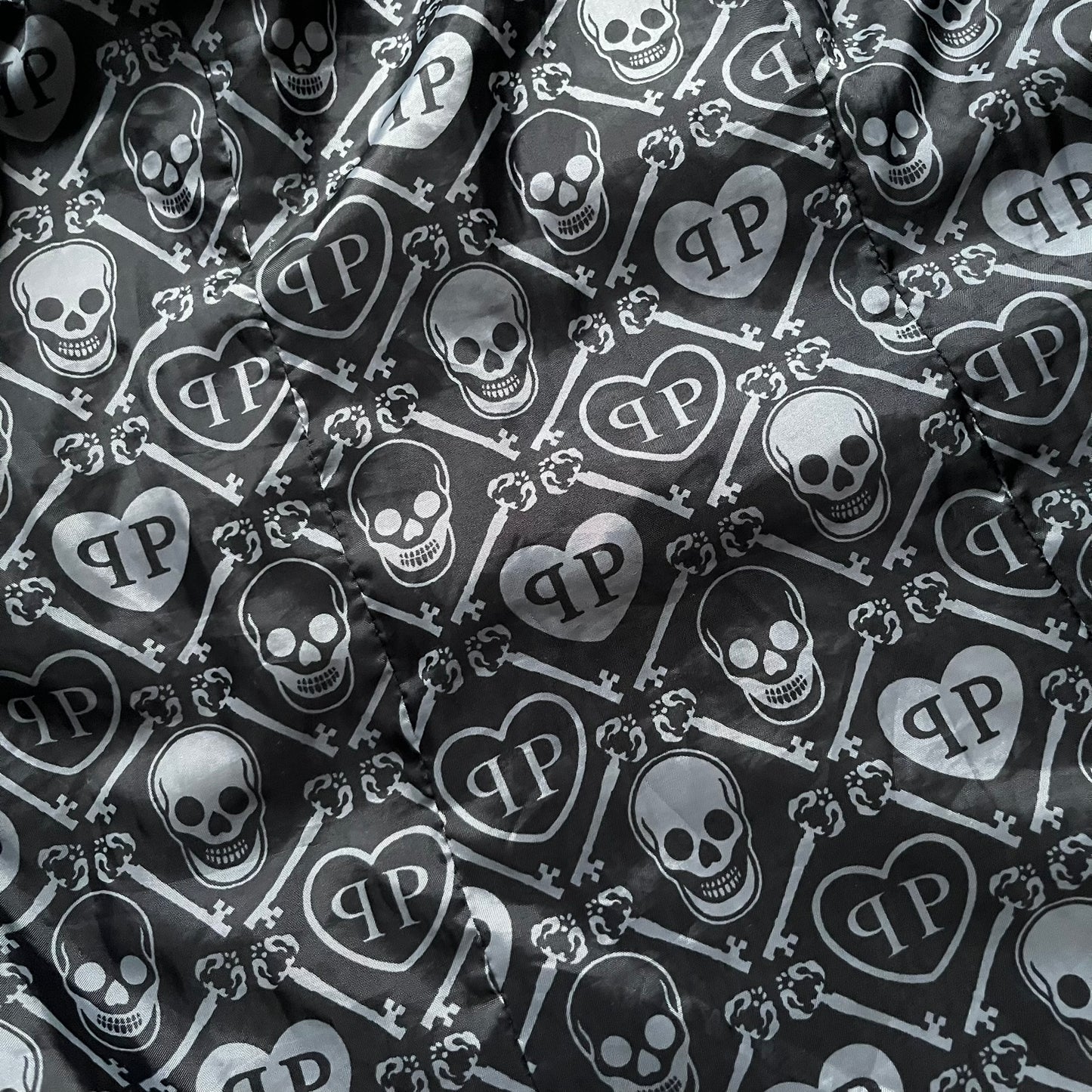 Philipp Plein 2010's No Hood Puffer Jacket is a black puffer jacket, with silver-tone hardware throughout including large zipper pulls shaped as crosses with a skull in the center used on the pockets, buttons with PP Skulls and metal "spikes" on the shoulders. The right arm has a white/clear “P” patch set with crystals. The interior lining has an all-over print featuring keys, skulls, and PP logos.