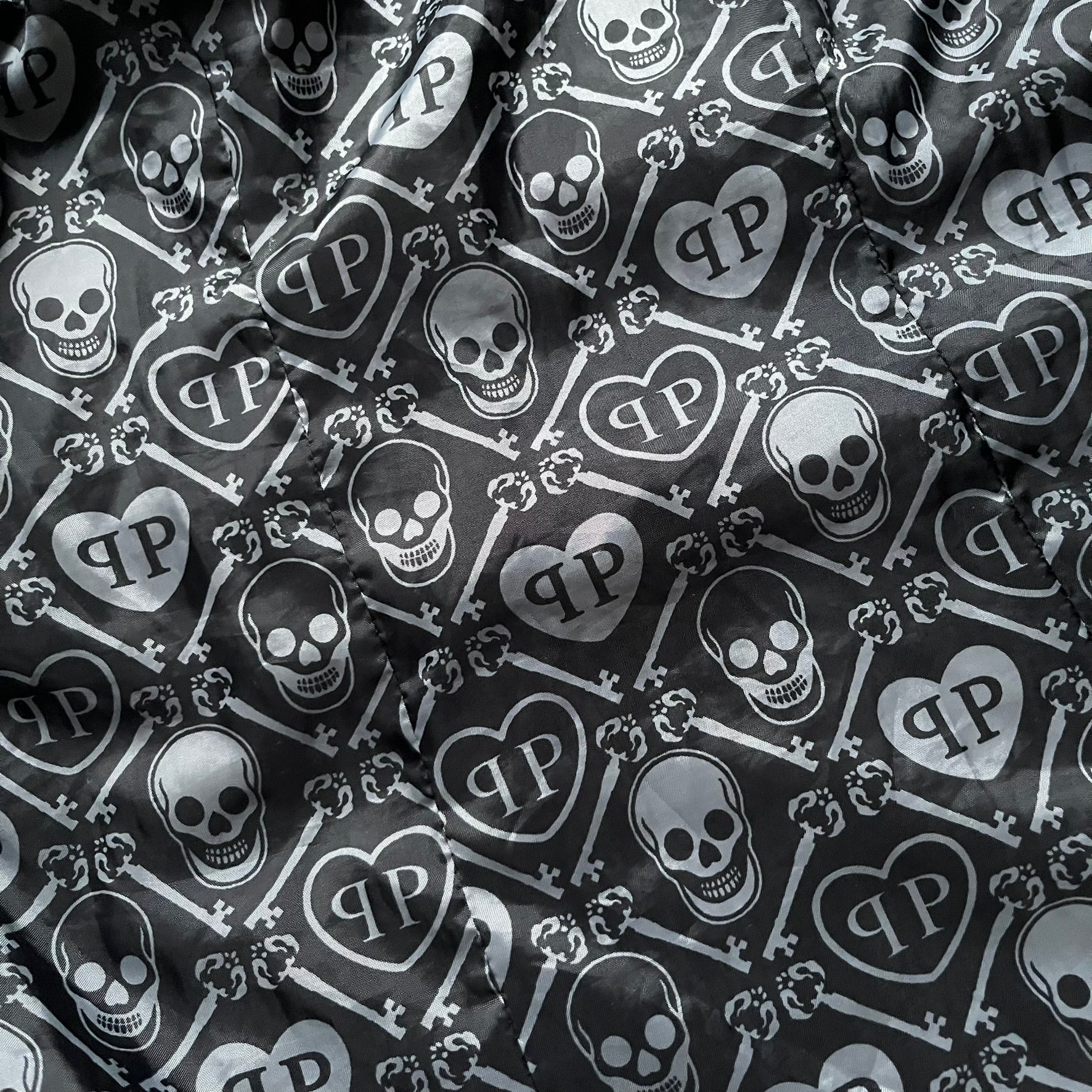 Philipp Plein 2010's No Hood Puffer Jacket is a black puffer jacket, with silver-tone hardware throughout including large zipper pulls shaped as crosses with a skull in the center used on the pockets, buttons with PP Skulls and metal "spikes" on the shoulders. The right arm has a white/clear “P” patch set with crystals. The interior lining has an all-over print featuring keys, skulls, and PP logos.