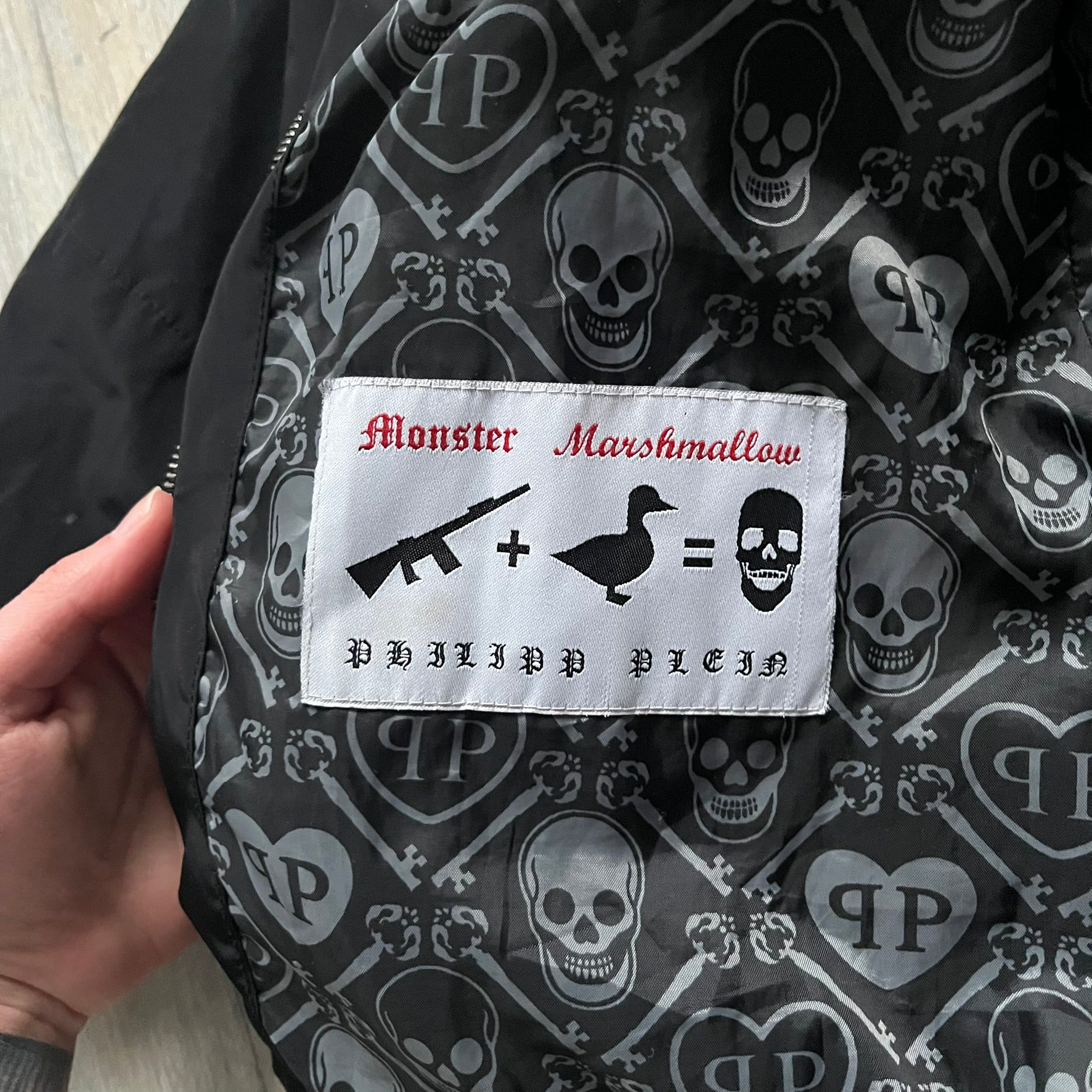 Philipp Plein 2010's No Hood Puffer Jacket is a black puffer jacket, with silver-tone hardware throughout including large zipper pulls shaped as crosses with a skull in the center used on the pockets, buttons with PP Skulls and metal "spikes" on the shoulders. The right arm has a white/clear “P” patch set with crystals. The interior lining has an all-over print featuring keys, skulls, and PP logos.