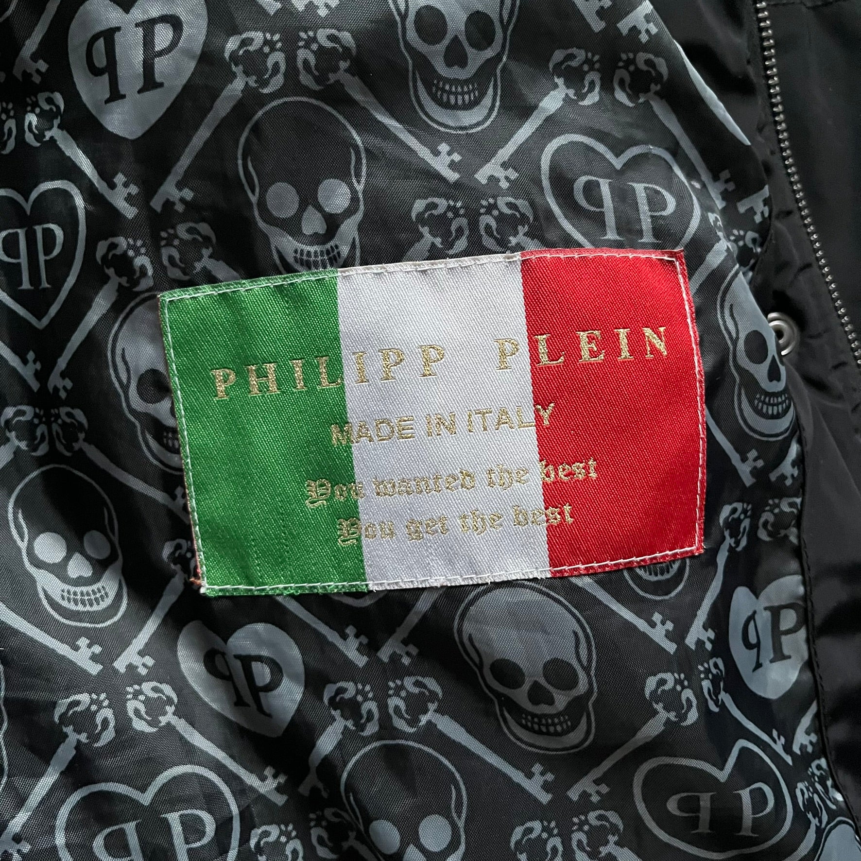 Philipp Plein 2010's No Hood Puffer Jacket is a black puffer jacket, with silver-tone hardware throughout including large zipper pulls shaped as crosses with a skull in the center used on the pockets, buttons with PP Skulls and metal "spikes" on the shoulders. The right arm has a white/clear “P” patch set with crystals. The interior lining has an all-over print featuring keys, skulls, and PP logos.
