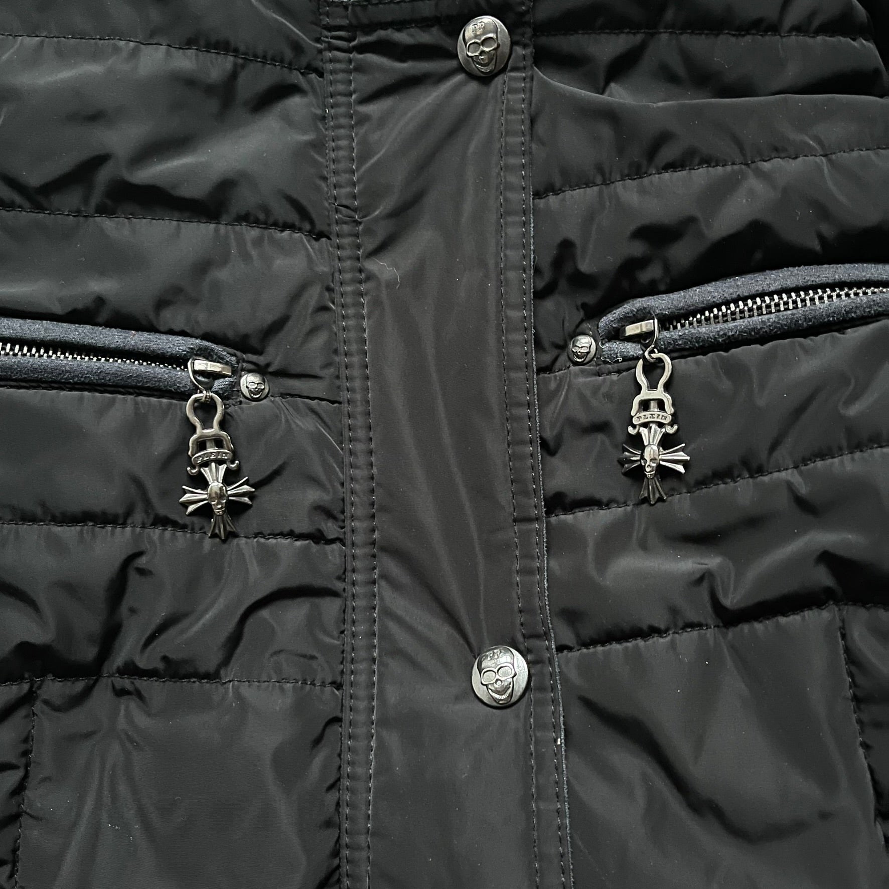 Philipp Plein 2010's No Hood Puffer Jacket is a black puffer jacket, with silver-tone hardware throughout including large zipper pulls shaped as crosses with a skull in the center used on the pockets, buttons with PP Skulls and metal "spikes" on the shoulders. The right arm has a white/clear “P” patch set with crystals. The interior lining has an all-over print featuring keys, skulls, and PP logos.