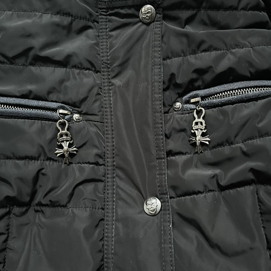 Philipp Plein 2010's No Hood Puffer Jacket is a black puffer jacket, with silver-tone hardware throughout including large zipper pulls shaped as crosses with a skull in the center used on the pockets, buttons with PP Skulls and metal "spikes" on the shoulders. The right arm has a white/clear “P” patch set with crystals. The interior lining has an all-over print featuring keys, skulls, and PP logos.