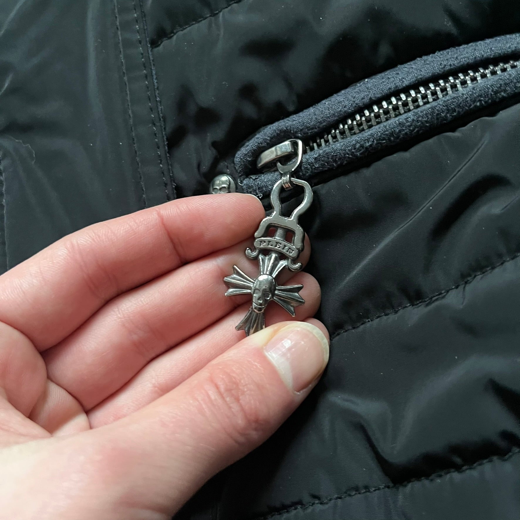Philipp Plein 2010's No Hood Puffer Jacket is a black puffer jacket, with silver-tone hardware throughout including large zipper pulls shaped as crosses with a skull in the center used on the pockets, buttons with PP Skulls and metal "spikes" on the shoulders. The right arm has a white/clear “P” patch set with crystals. The interior lining has an all-over print featuring keys, skulls, and PP logos.