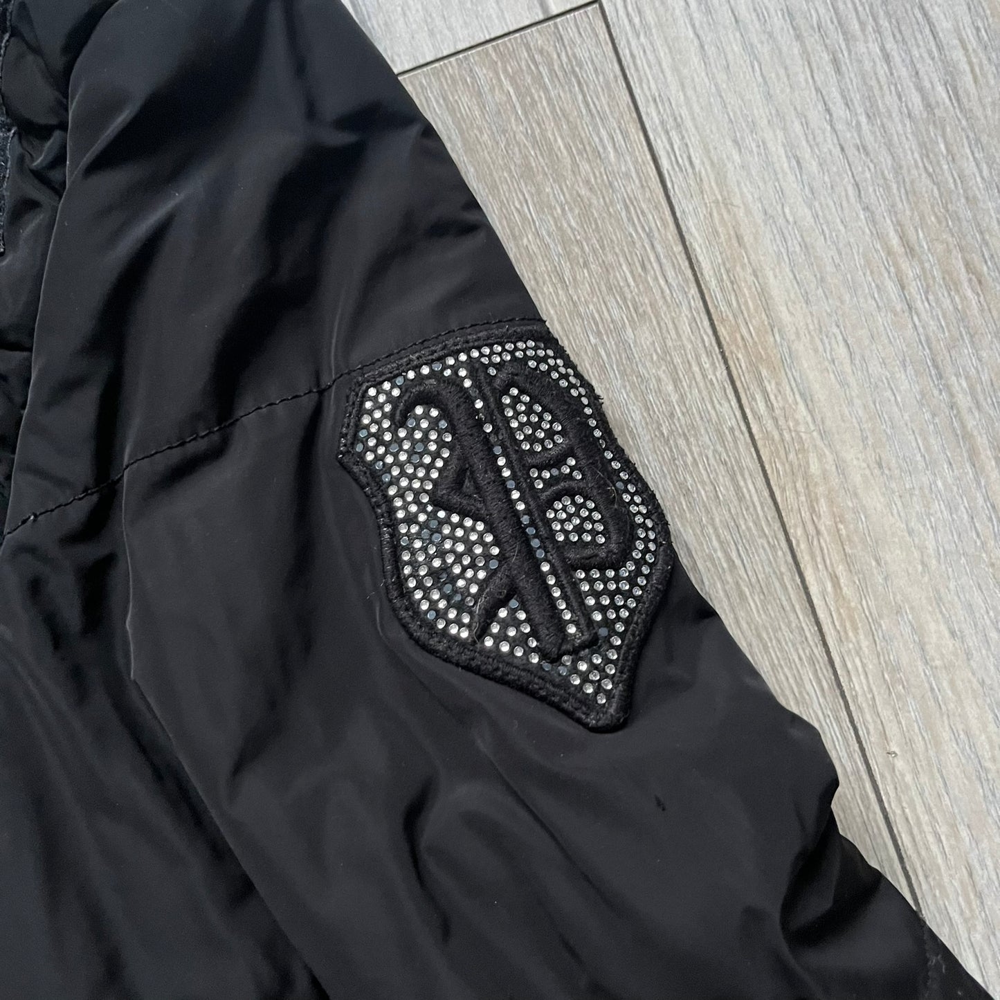 Philipp Plein 2010's No Hood Puffer Jacket is a black puffer jacket, with silver-tone hardware throughout including large zipper pulls shaped as crosses with a skull in the center used on the pockets, buttons with PP Skulls and metal "spikes" on the shoulders. The right arm has a white/clear “P” patch set with crystals. The interior lining has an all-over print featuring keys, skulls, and PP logos.