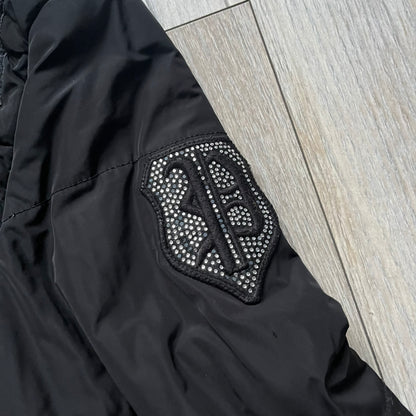 Philipp Plein 2010's No Hood Puffer Jacket is a black puffer jacket, with silver-tone hardware throughout including large zipper pulls shaped as crosses with a skull in the center used on the pockets, buttons with PP Skulls and metal "spikes" on the shoulders. The right arm has a white/clear “P” patch set with crystals. The interior lining has an all-over print featuring keys, skulls, and PP logos.