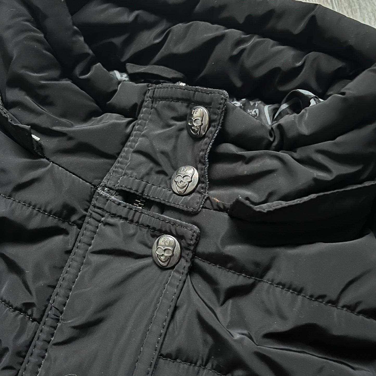 Philipp Plein 2010's No Hood Puffer Jacket is a black puffer jacket, with silver-tone hardware throughout including large zipper pulls shaped as crosses with a skull in the center used on the pockets, buttons with PP Skulls and metal "spikes" on the shoulders. The right arm has a white/clear “P” patch set with crystals. The interior lining has an all-over print featuring keys, skulls, and PP logos.