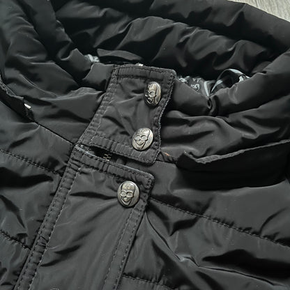 Philipp Plein 2010's No Hood Puffer Jacket is a black puffer jacket, with silver-tone hardware throughout including large zipper pulls shaped as crosses with a skull in the center used on the pockets, buttons with PP Skulls and metal "spikes" on the shoulders. The right arm has a white/clear “P” patch set with crystals. The interior lining has an all-over print featuring keys, skulls, and PP logos.