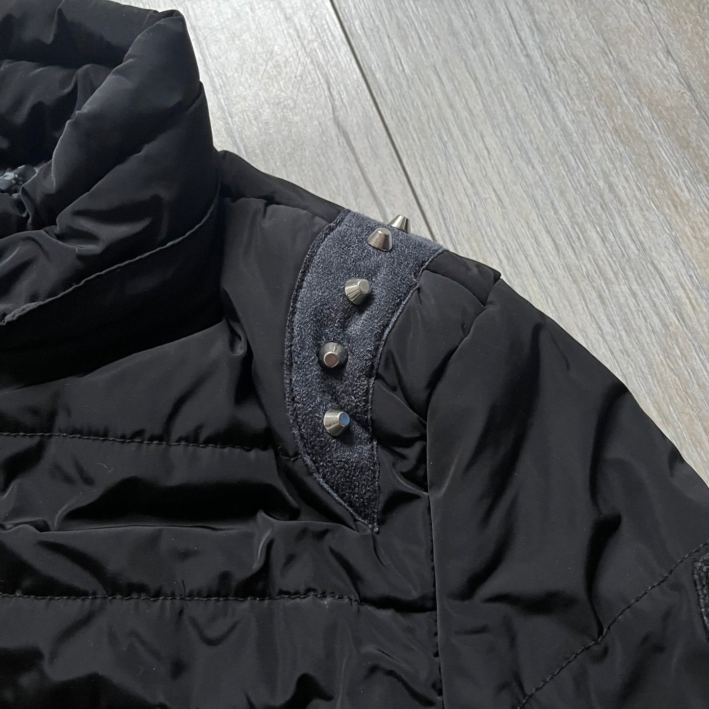 Philipp Plein 2010's No Hood Puffer Jacket is a black puffer jacket, with silver-tone hardware throughout including large zipper pulls shaped as crosses with a skull in the center used on the pockets, buttons with PP Skulls and metal "spikes" on the shoulders. The right arm has a white/clear “P” patch set with crystals. The interior lining has an all-over print featuring keys, skulls, and PP logos.