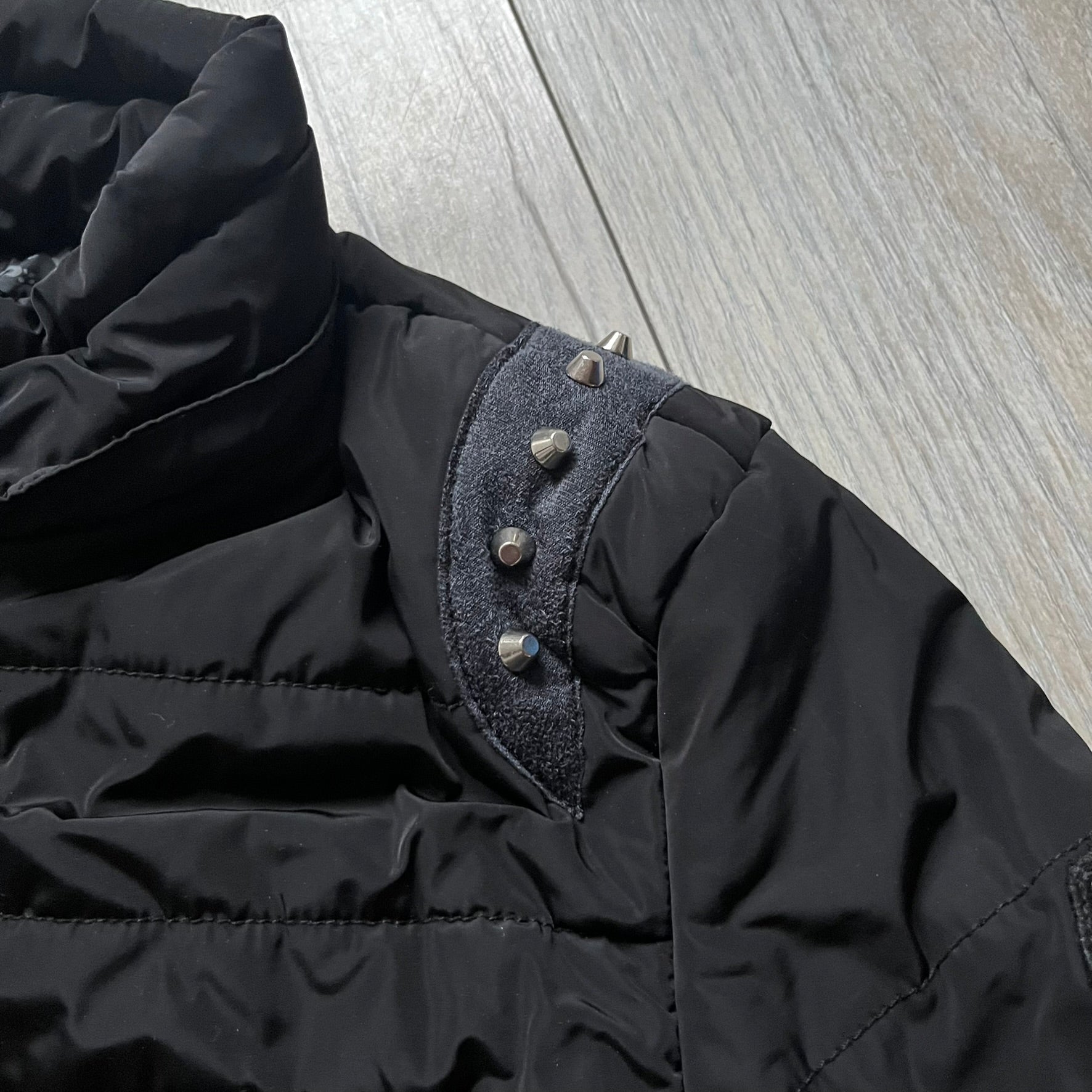 Philipp Plein 2010's No Hood Puffer Jacket is a black puffer jacket, with silver-tone hardware throughout including large zipper pulls shaped as crosses with a skull in the center used on the pockets, buttons with PP Skulls and metal "spikes" on the shoulders. The right arm has a white/clear “P” patch set with crystals. The interior lining has an all-over print featuring keys, skulls, and PP logos.