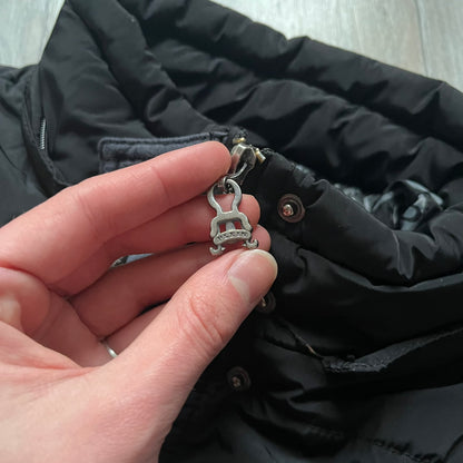 Philipp Plein 2010's No Hood Puffer Jacket is a black puffer jacket, with silver-tone hardware throughout including large zipper pulls shaped as crosses with a skull in the center used on the pockets, buttons with PP Skulls and metal "spikes" on the shoulders. The right arm has a white/clear “P” patch set with crystals. The interior lining has an all-over print featuring keys, skulls, and PP logos.