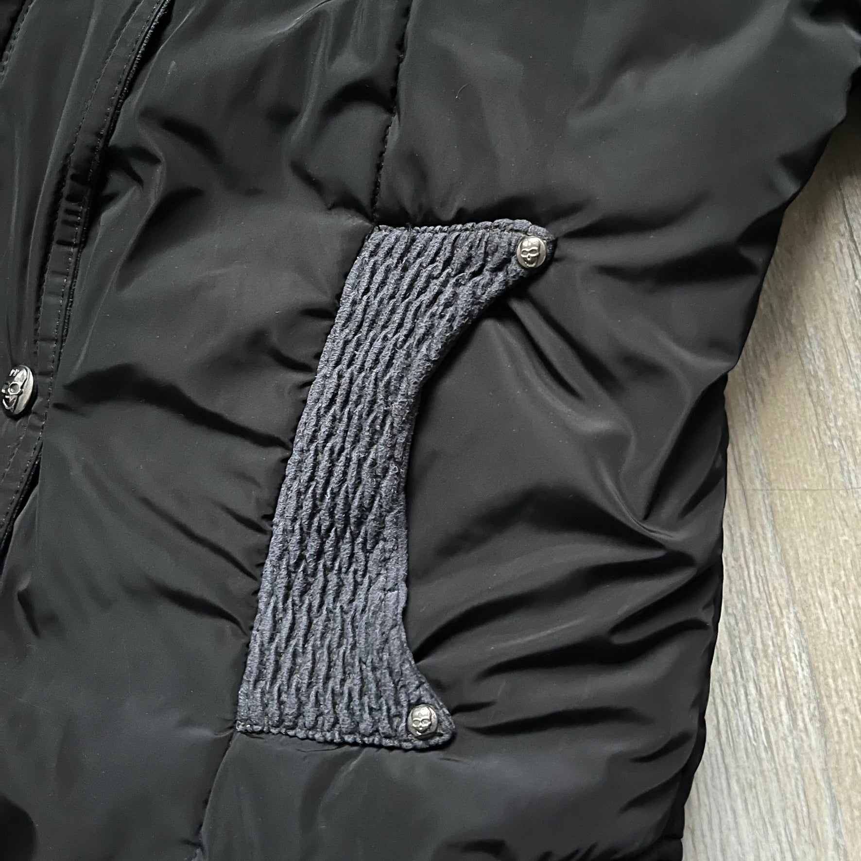 Philipp Plein 2010's No Hood Puffer Jacket is a black puffer jacket, with silver-tone hardware throughout including large zipper pulls shaped as crosses with a skull in the center used on the pockets, buttons with PP Skulls and metal "spikes" on the shoulders. The right arm has a white/clear “P” patch set with crystals. The interior lining has an all-over print featuring keys, skulls, and PP logos.