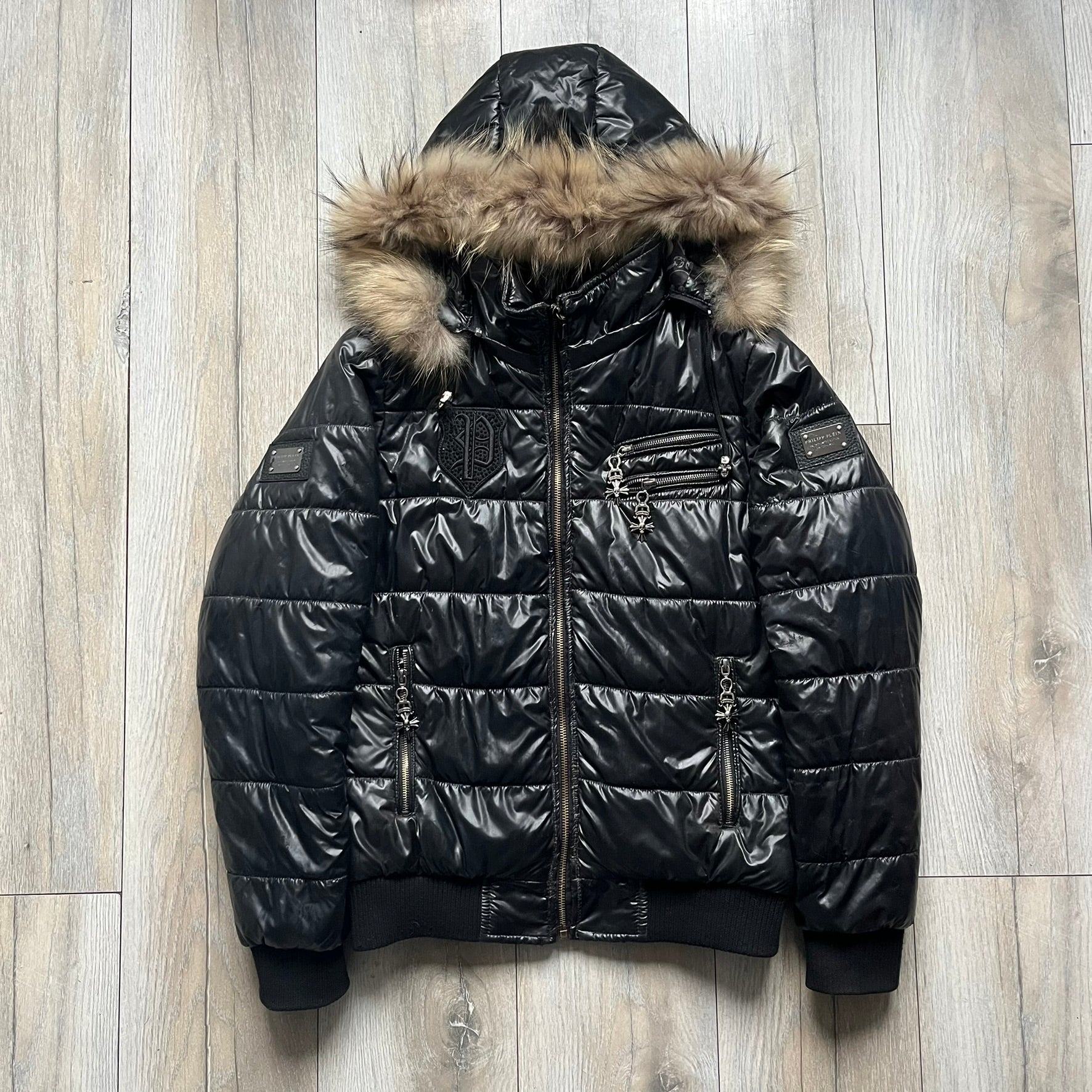 Philipp Plein 2010s Black Waxed Puffer Jacket is a black puffer jacket with a removable fur-trimmed hood attached by zipper. The jacket features heavy silver-tone hardware throughout, including large zipper pulls shaped as crosses with a skull in the center, used on the front pockets and chest pockets. The right chest has a black “P” patch set with crystals, while the left chest features two zip pockets. The hood drawstrings are finished with silver skull tips marked with a “P.”

Both shoulders are finished