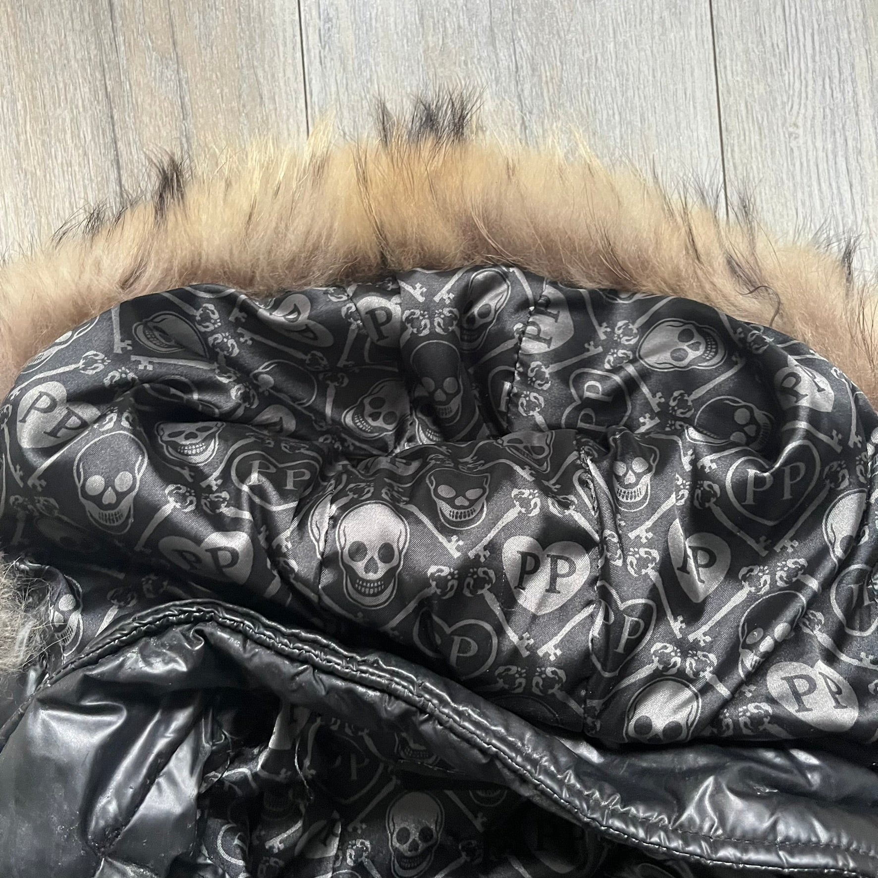 Philipp Plein 2010s Black Waxed Puffer Jacket is a black puffer jacket with a removable fur-trimmed hood attached by zipper. The jacket features heavy silver-tone hardware throughout, including large zipper pulls shaped as crosses with a skull in the center, used on the front pockets and chest pockets. The right chest has a black “P” patch set with crystals, while the left chest features two zip pockets. The hood drawstrings are finished with silver skull tips marked with a “P.”

Both shoulders are finished