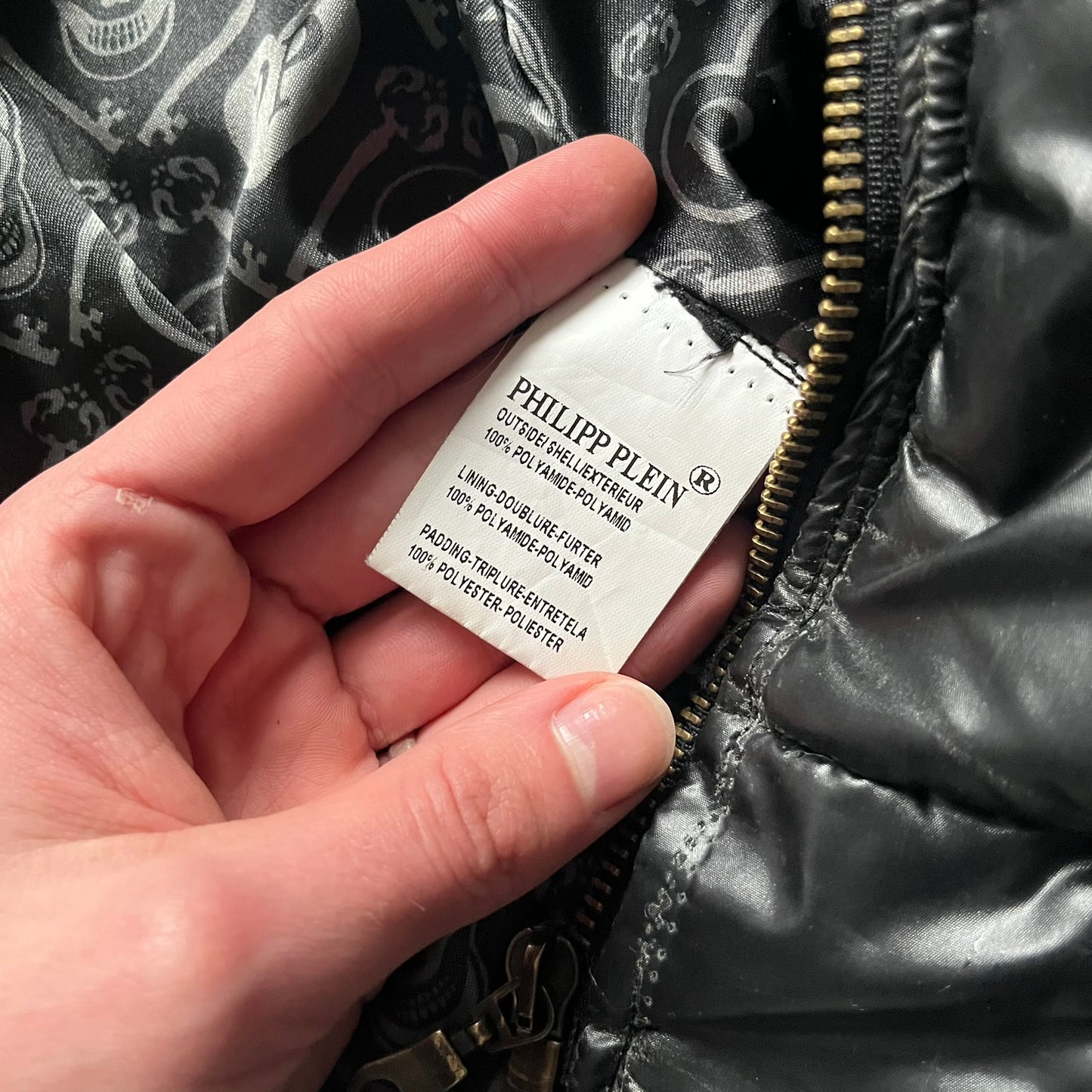 Philipp Plein 2010s Black Waxed Puffer Jacket is a black puffer jacket with a removable fur-trimmed hood attached by zipper. The jacket features heavy silver-tone hardware throughout, including large zipper pulls shaped as crosses with a skull in the center, used on the front pockets and chest pockets. The right chest has a black “P” patch set with crystals, while the left chest features two zip pockets. The hood drawstrings are finished with silver skull tips marked with a “P.”

Both shoulders are finished