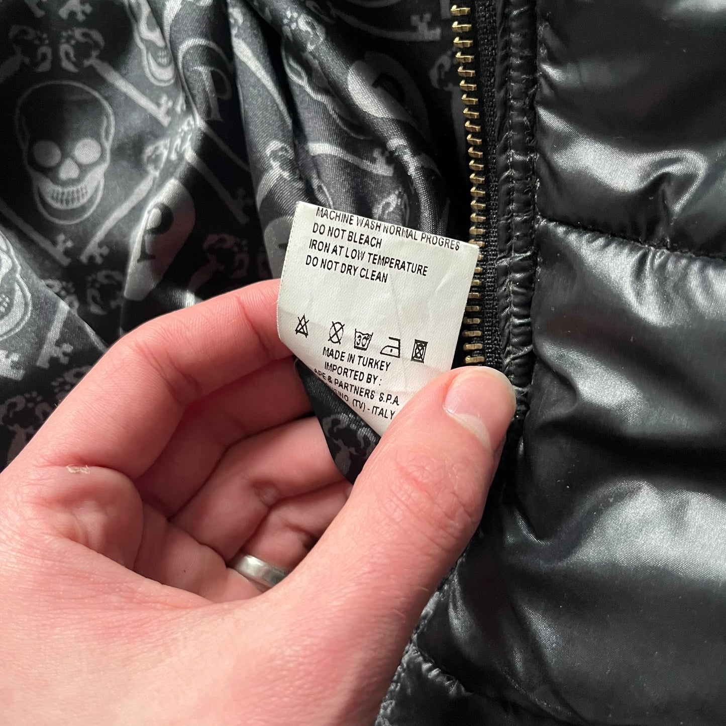 Philipp Plein 2010s Black Waxed Puffer Jacket is a black puffer jacket with a removable fur-trimmed hood attached by zipper. The jacket features heavy silver-tone hardware throughout, including large zipper pulls shaped as crosses with a skull in the center, used on the front pockets and chest pockets. The right chest has a black “P” patch set with crystals, while the left chest features two zip pockets. The hood drawstrings are finished with silver skull tips marked with a “P.”

Both shoulders are finished