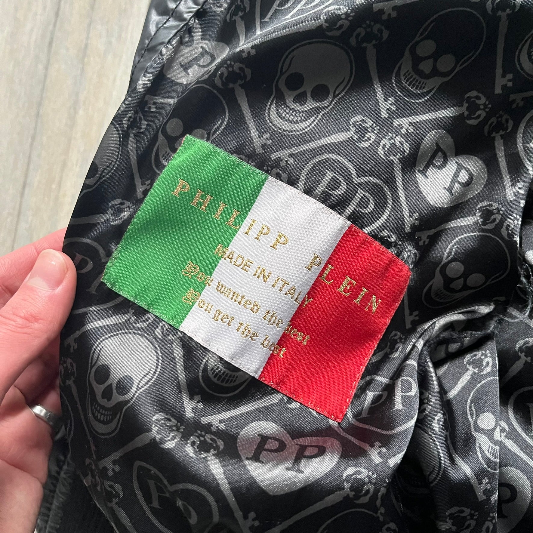 Philipp Plein 2010s Black Waxed Puffer Jacket is a black puffer jacket with a removable fur-trimmed hood attached by zipper. The jacket features heavy silver-tone hardware throughout, including large zipper pulls shaped as crosses with a skull in the center, used on the front pockets and chest pockets. The right chest has a black “P” patch set with crystals, while the left chest features two zip pockets. The hood drawstrings are finished with silver skull tips marked with a “P.”

Both shoulders are finished