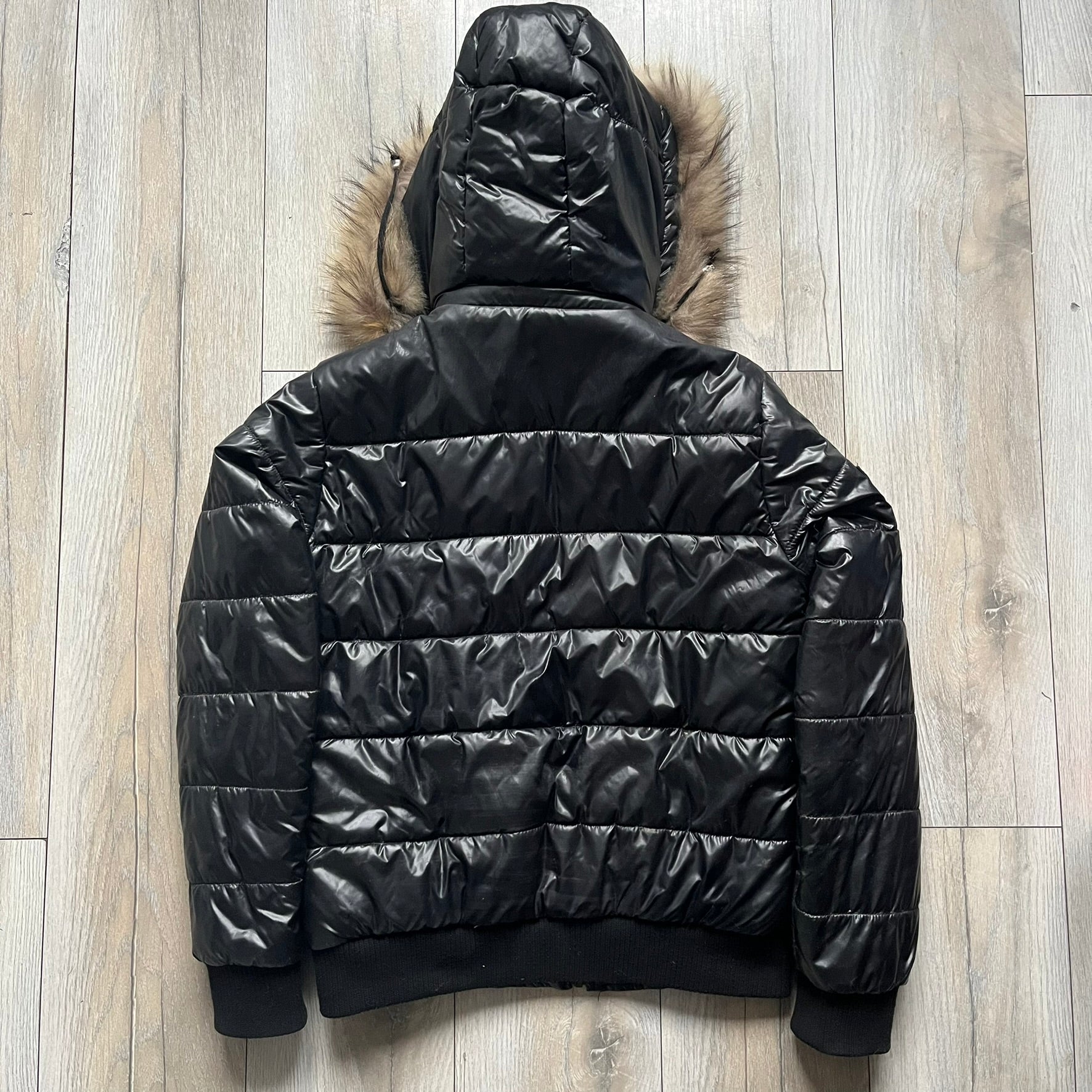 Philipp Plein 2010s Black Waxed Puffer Jacket is a black puffer jacket with a removable fur-trimmed hood attached by zipper. The jacket features heavy silver-tone hardware throughout, including large zipper pulls shaped as crosses with a skull in the center, used on the front pockets and chest pockets. The right chest has a black “P” patch set with crystals, while the left chest features two zip pockets. The hood drawstrings are finished with silver skull tips marked with a “P.”

Both shoulders are finished