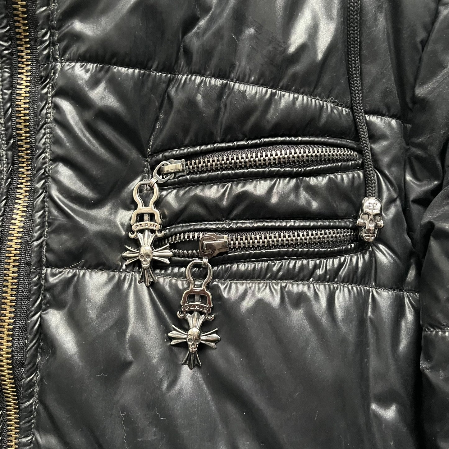 Philipp Plein 2010s Black Waxed Puffer Jacket is a black puffer jacket with a removable fur-trimmed hood attached by zipper. The jacket features heavy silver-tone hardware throughout, including large zipper pulls shaped as crosses with a skull in the center, used on the front pockets and chest pockets. The right chest has a black “P” patch set with crystals, while the left chest features two zip pockets. The hood drawstrings are finished with silver skull tips marked with a “P.”

Both shoulders are finished