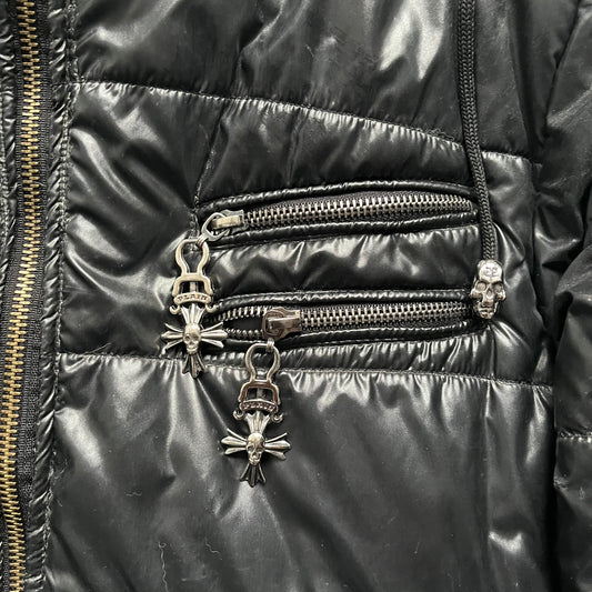 Philipp Plein 2010s Black Waxed Puffer Jacket is a black puffer jacket with a removable fur-trimmed hood attached by zipper. The jacket features heavy silver-tone hardware throughout, including large zipper pulls shaped as crosses with a skull in the center, used on the front pockets and chest pockets. The right chest has a black “P” patch set with crystals, while the left chest features two zip pockets. The hood drawstrings are finished with silver skull tips marked with a “P.”

Both shoulders are finished