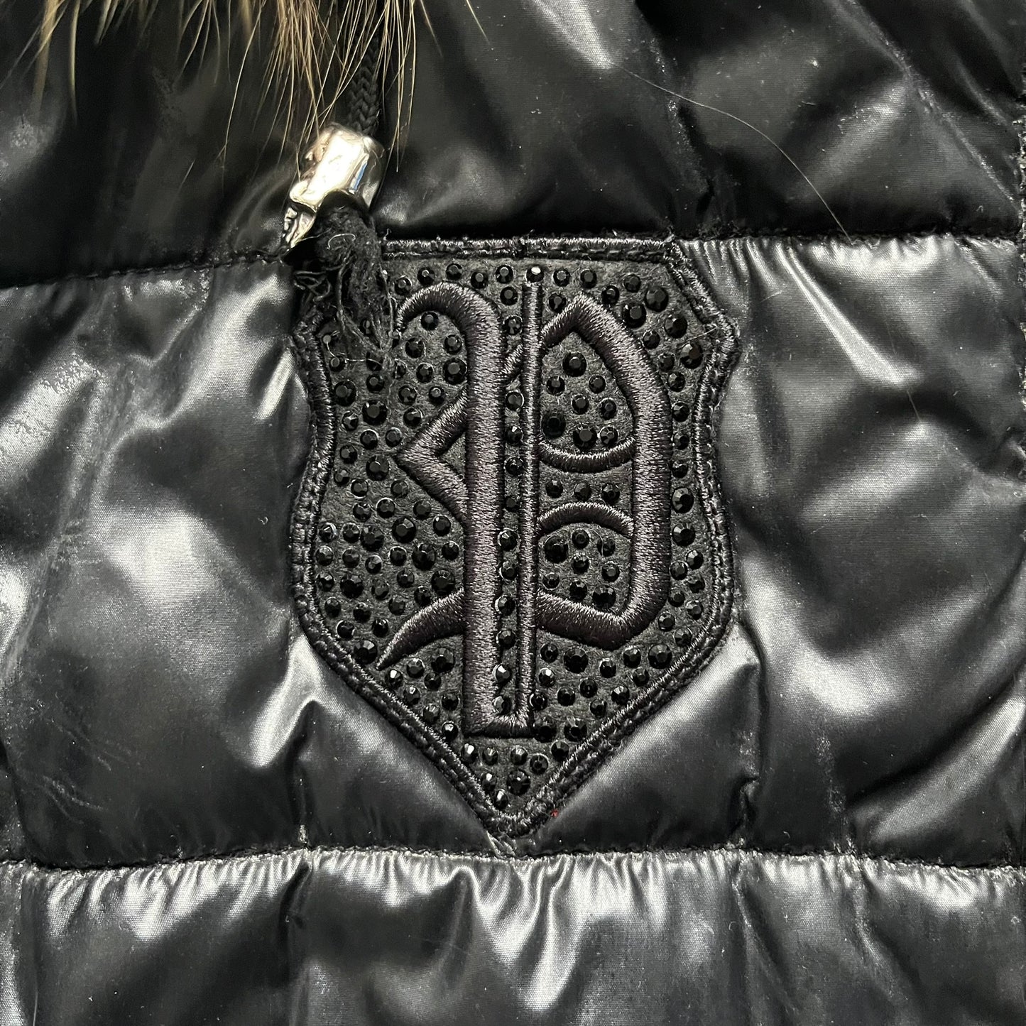 Philipp Plein 2010s Black Waxed Puffer Jacket is a black puffer jacket with a removable fur-trimmed hood attached by zipper. The jacket features heavy silver-tone hardware throughout, including large zipper pulls shaped as crosses with a skull in the center, used on the front pockets and chest pockets. The right chest has a black “P” patch set with crystals, while the left chest features two zip pockets. The hood drawstrings are finished with silver skull tips marked with a “P.”

Both shoulders are finished