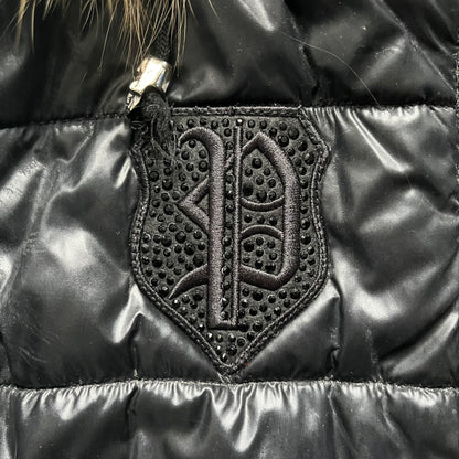 Philipp Plein 2010s Black Waxed Puffer Jacket is a black puffer jacket with a removable fur-trimmed hood attached by zipper. The jacket features heavy silver-tone hardware throughout, including large zipper pulls shaped as crosses with a skull in the center, used on the front pockets and chest pockets. The right chest has a black “P” patch set with crystals, while the left chest features two zip pockets. The hood drawstrings are finished with silver skull tips marked with a “P.”

Both shoulders are finished