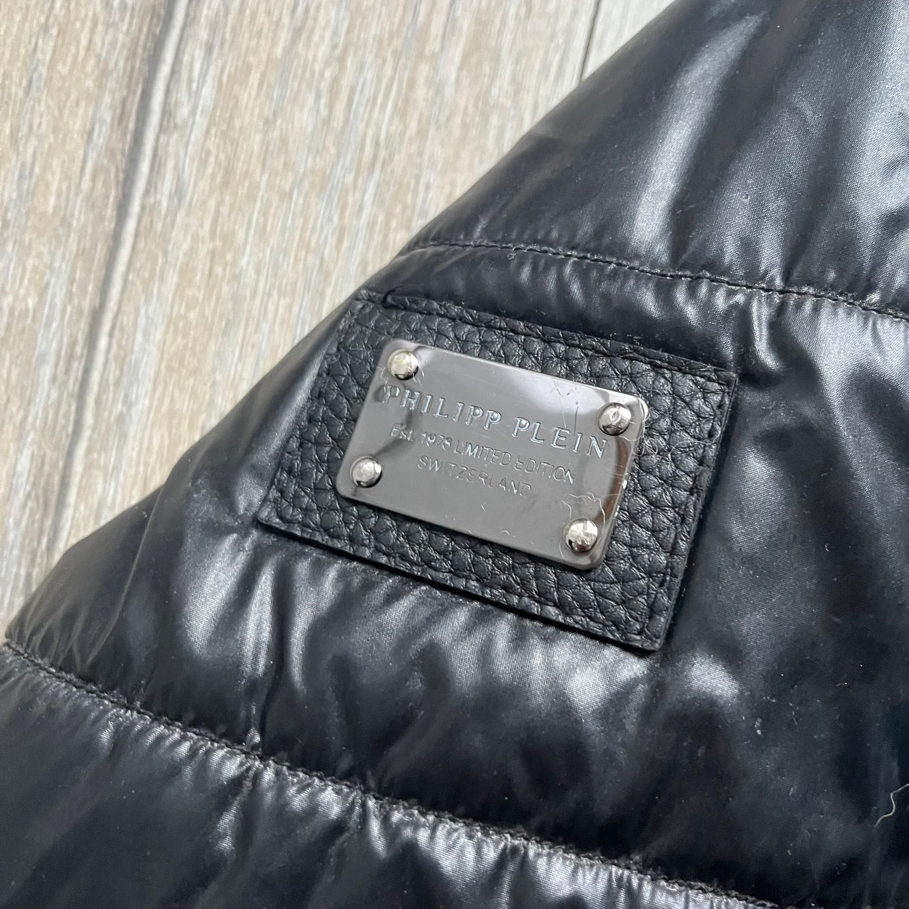 Philipp Plein 2010s Black Waxed Puffer Jacket is a black puffer jacket with a removable fur-trimmed hood attached by zipper. The jacket features heavy silver-tone hardware throughout, including large zipper pulls shaped as crosses with a skull in the center, used on the front pockets and chest pockets. The right chest has a black “P” patch set with crystals, while the left chest features two zip pockets. The hood drawstrings are finished with silver skull tips marked with a “P.”

Both shoulders are finished