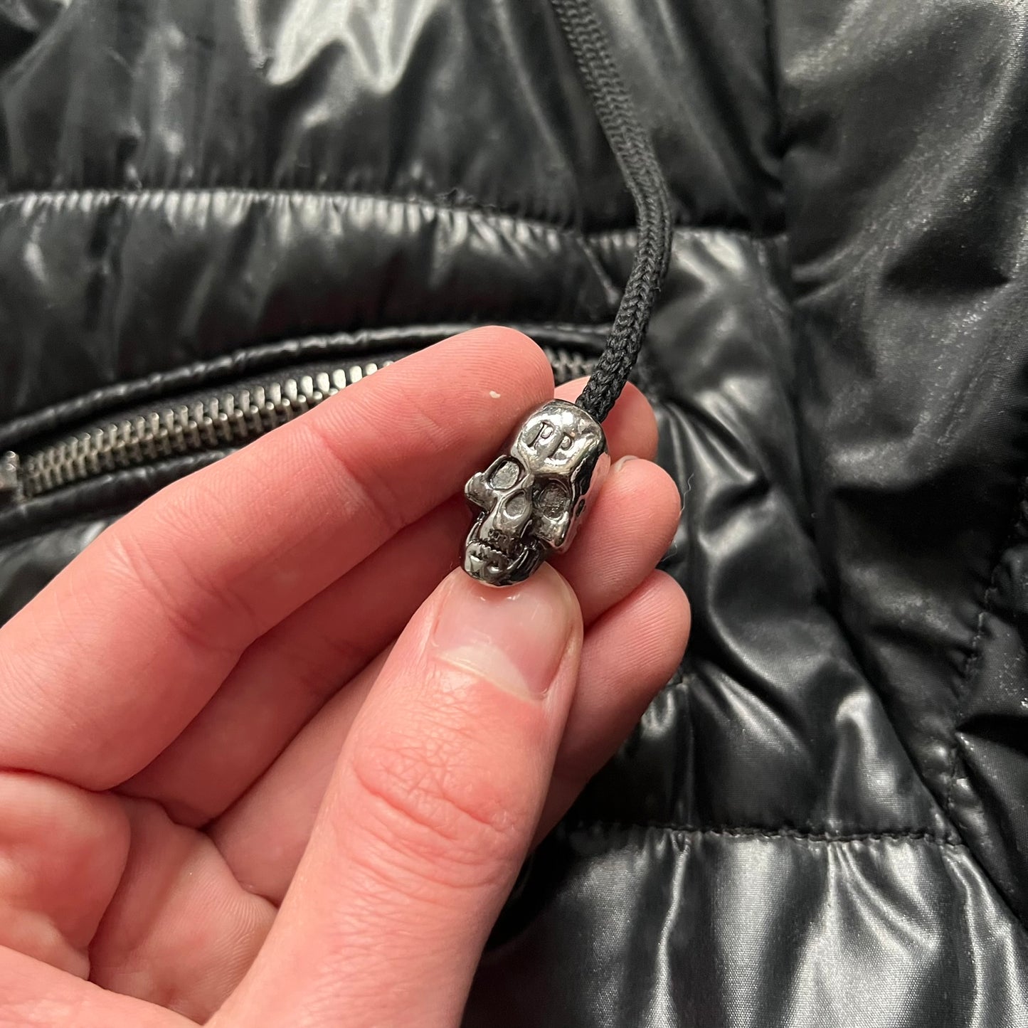 Philipp Plein 2010s Black Waxed Puffer Jacket is a black puffer jacket with a removable fur-trimmed hood attached by zipper. The jacket features heavy silver-tone hardware throughout, including large zipper pulls shaped as crosses with a skull in the center, used on the front pockets and chest pockets. The right chest has a black “P” patch set with crystals, while the left chest features two zip pockets. The hood drawstrings are finished with silver skull tips marked with a “P.”

Both shoulders are finished